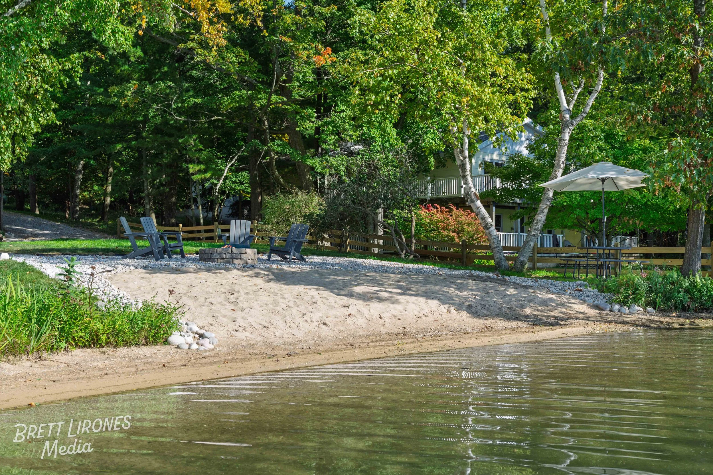 A lakeside scene with sandy shore, four Adirondack chairs arranged around a fire pit, and a patio with a table, chairs, and umbrella, surrounded by lush green trees and residential buildings in the background.