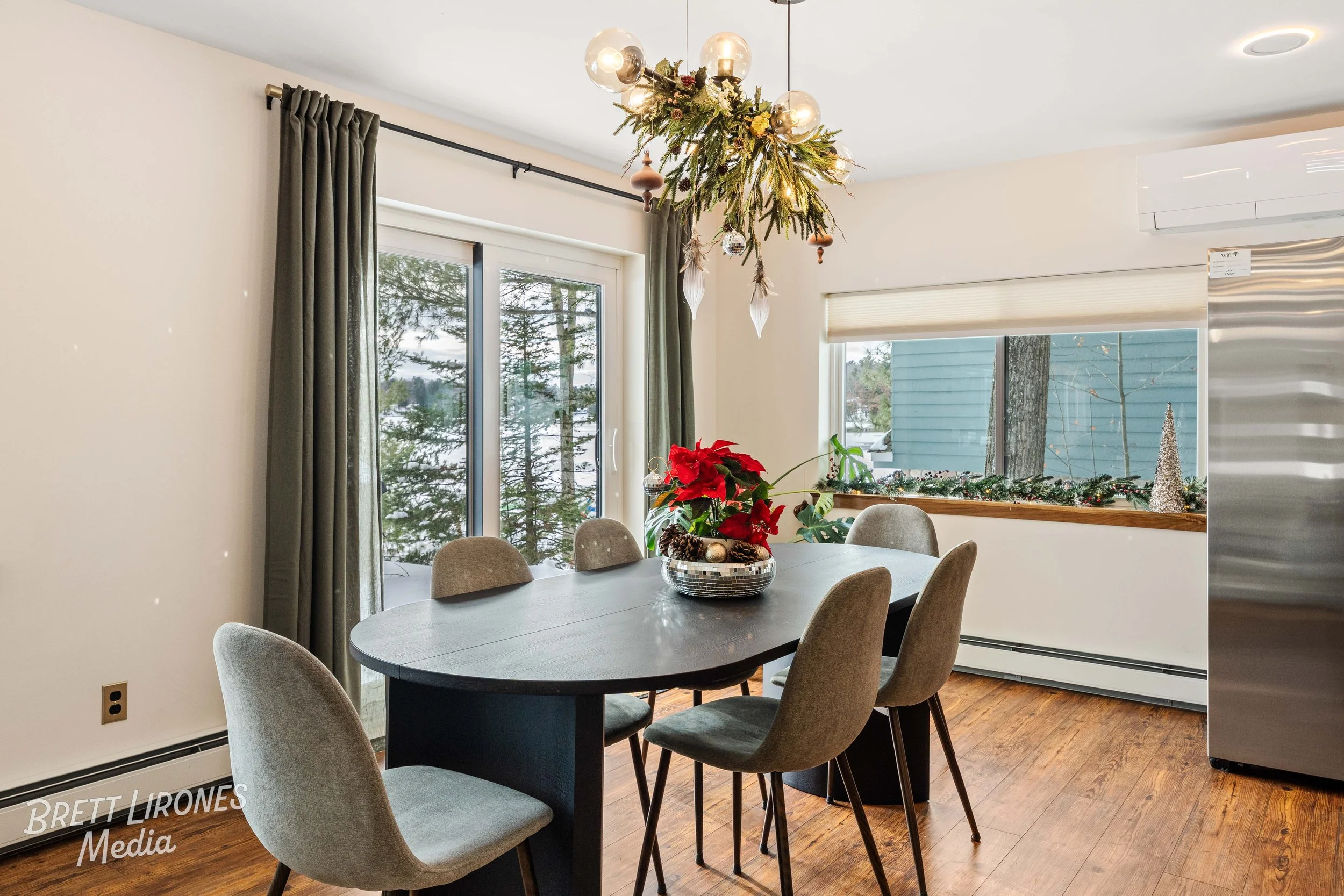 Dining room with a black table, six beige chairs, a poinsettia centerpiece, silver and glass decorations, large window with snow outside, and a festive garland and cones on the window sill, decorated for Christmas.