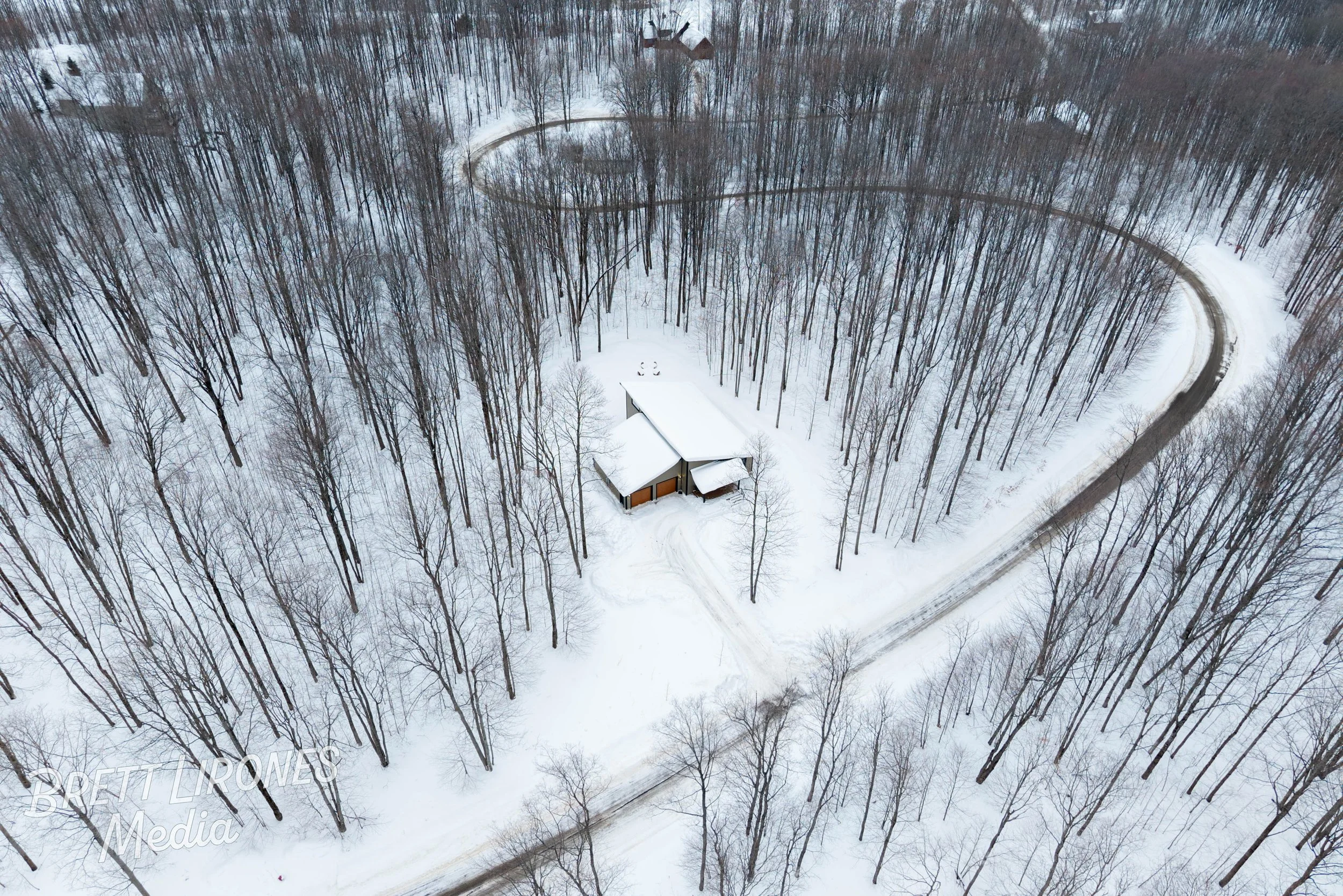 A snow-covered landscape with a house and garages surrounded by leafless trees and a winding road in winter.
