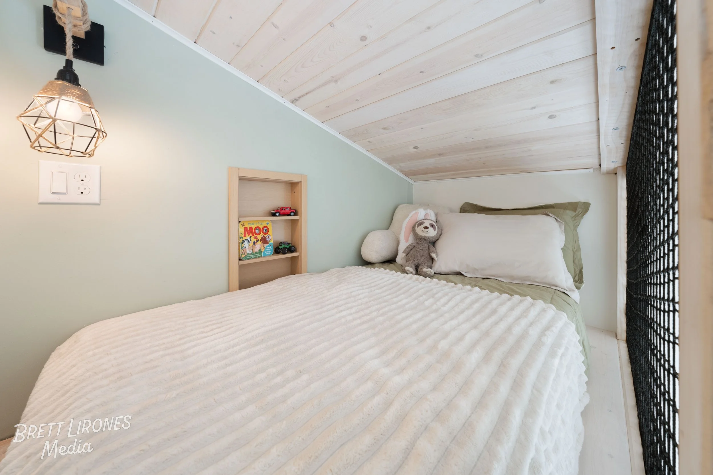 Cozy bedroom with a sloped wooden ceiling, white and green bedding, stuffed animals including a plush bunny and a sloth, a small wooden shelf with toy cars and a book, and a geometric wall lamp.