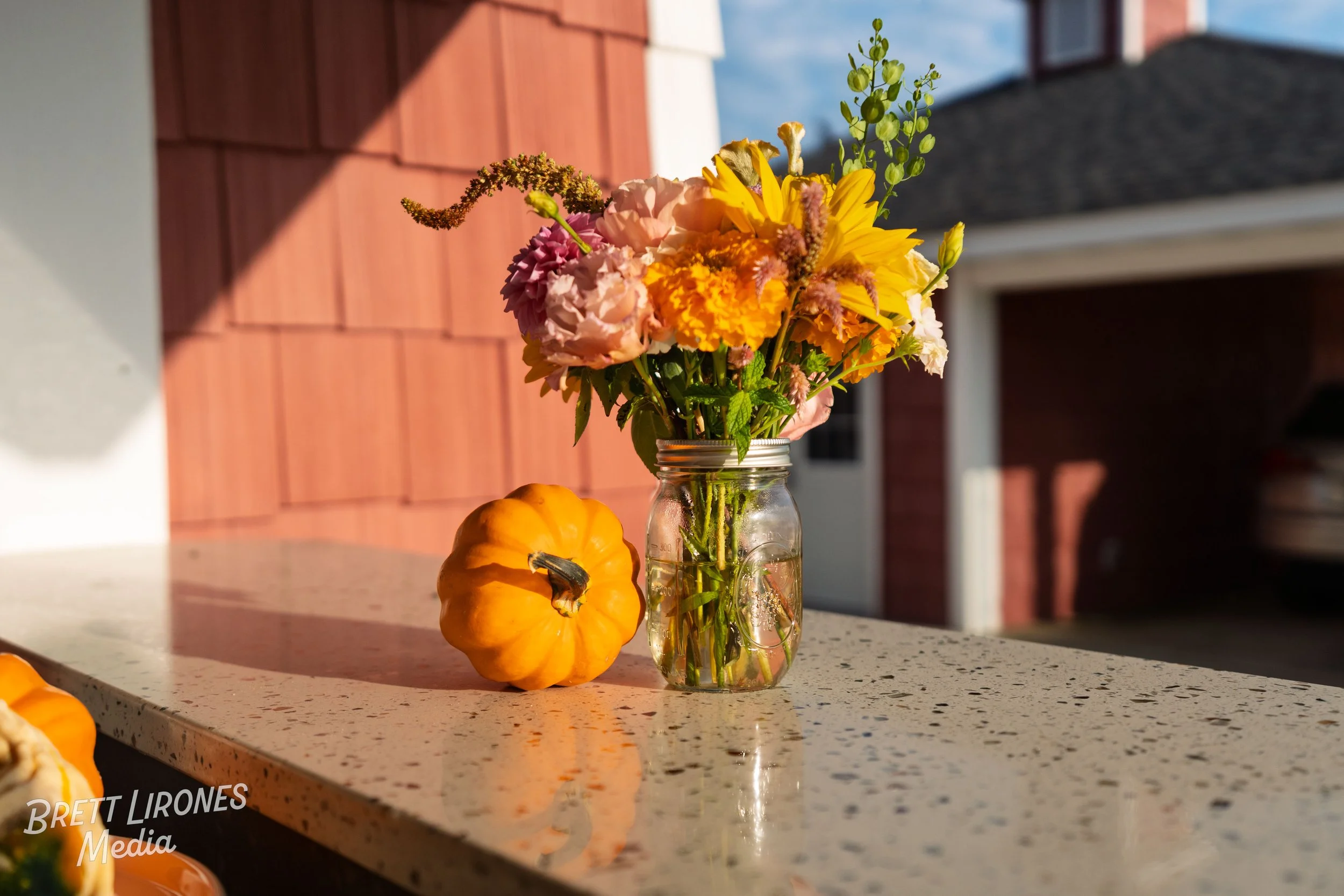 A mason jar filled with colorful flowers on a speckled countertop, beside two small orange gourds, with a red wooden wall and a house in the background.