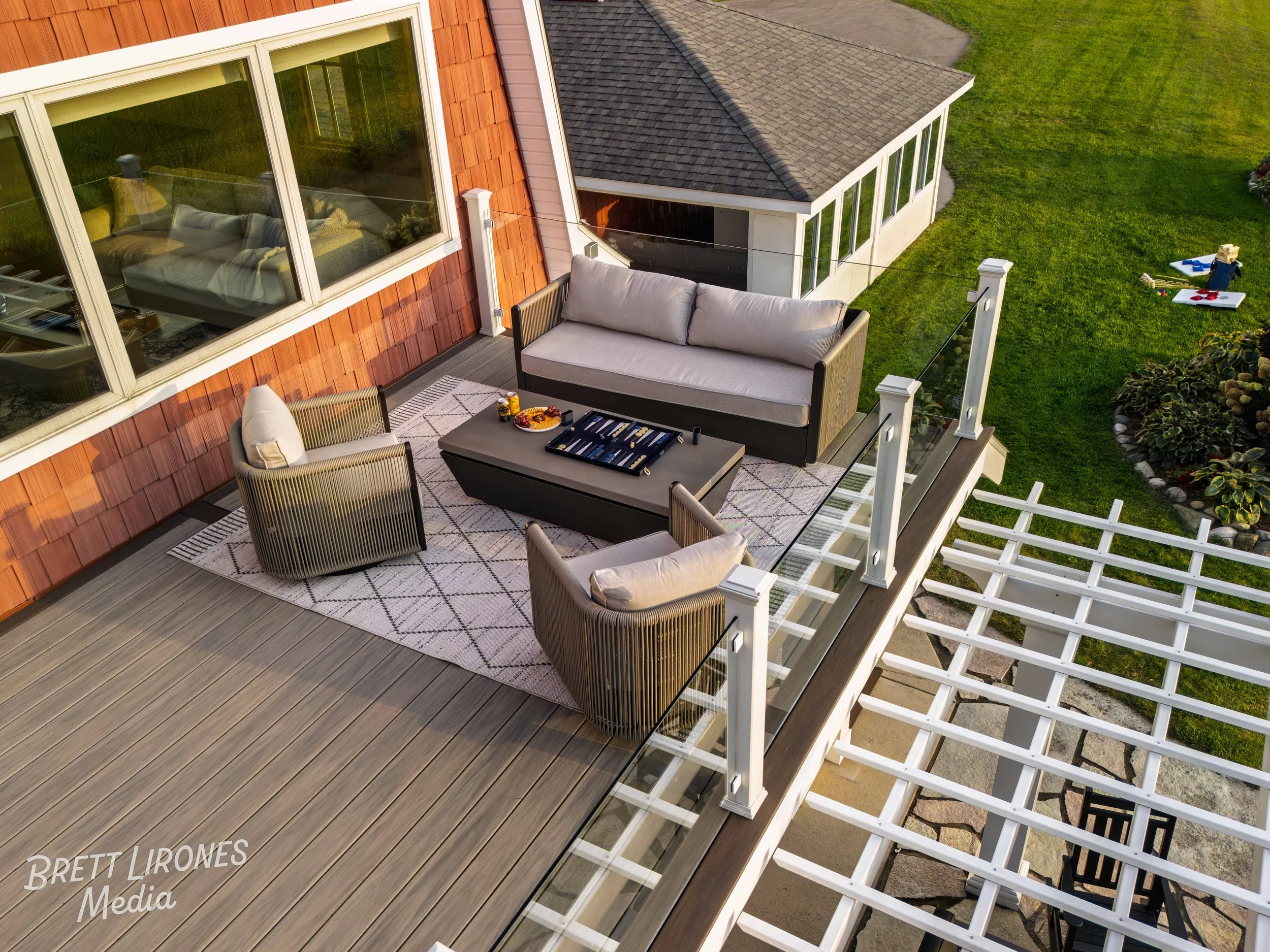 Outdoor balcony with seating area, including a cushioned loveseat and two armchairs around a coffee table, on a wooden deck with a rug, overlooking a grassy yard with children's toys