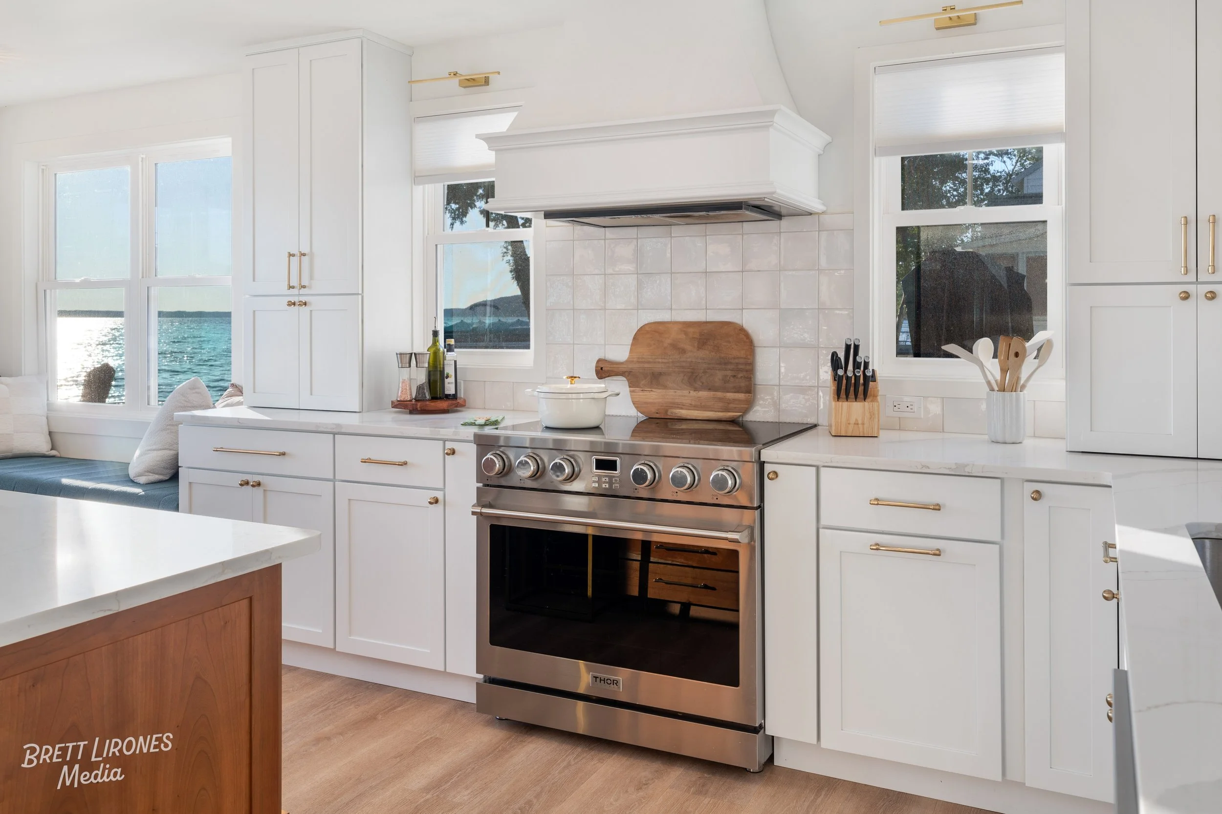 Modern kitchen with white cabinets and a stainless steel oven, near windows overlooking a body of water with mountains in the distance.
