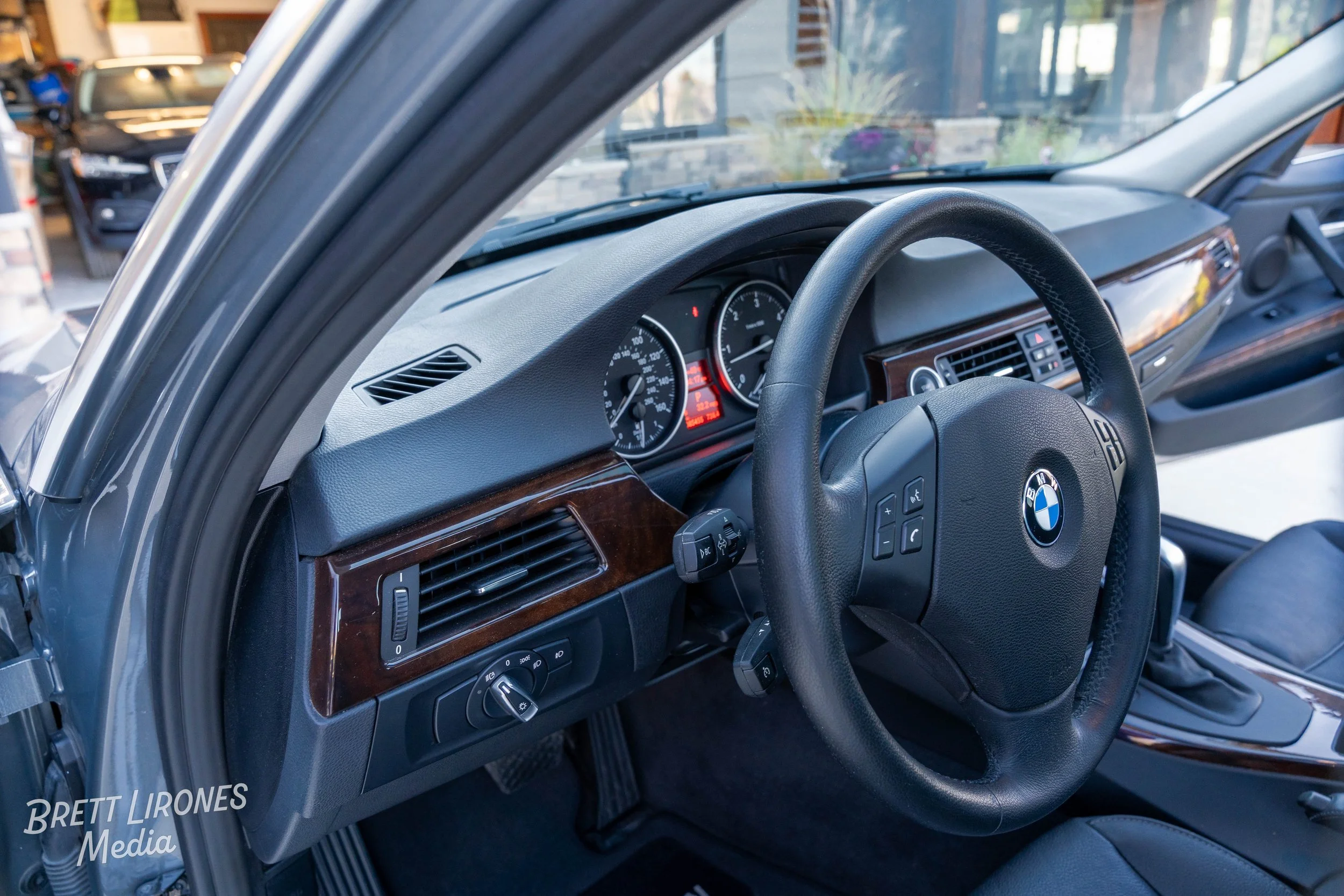 Interior of a BMW car showing the steering wheel and dashboard with wood trim, including instrument gauges, air vents, and control buttons.