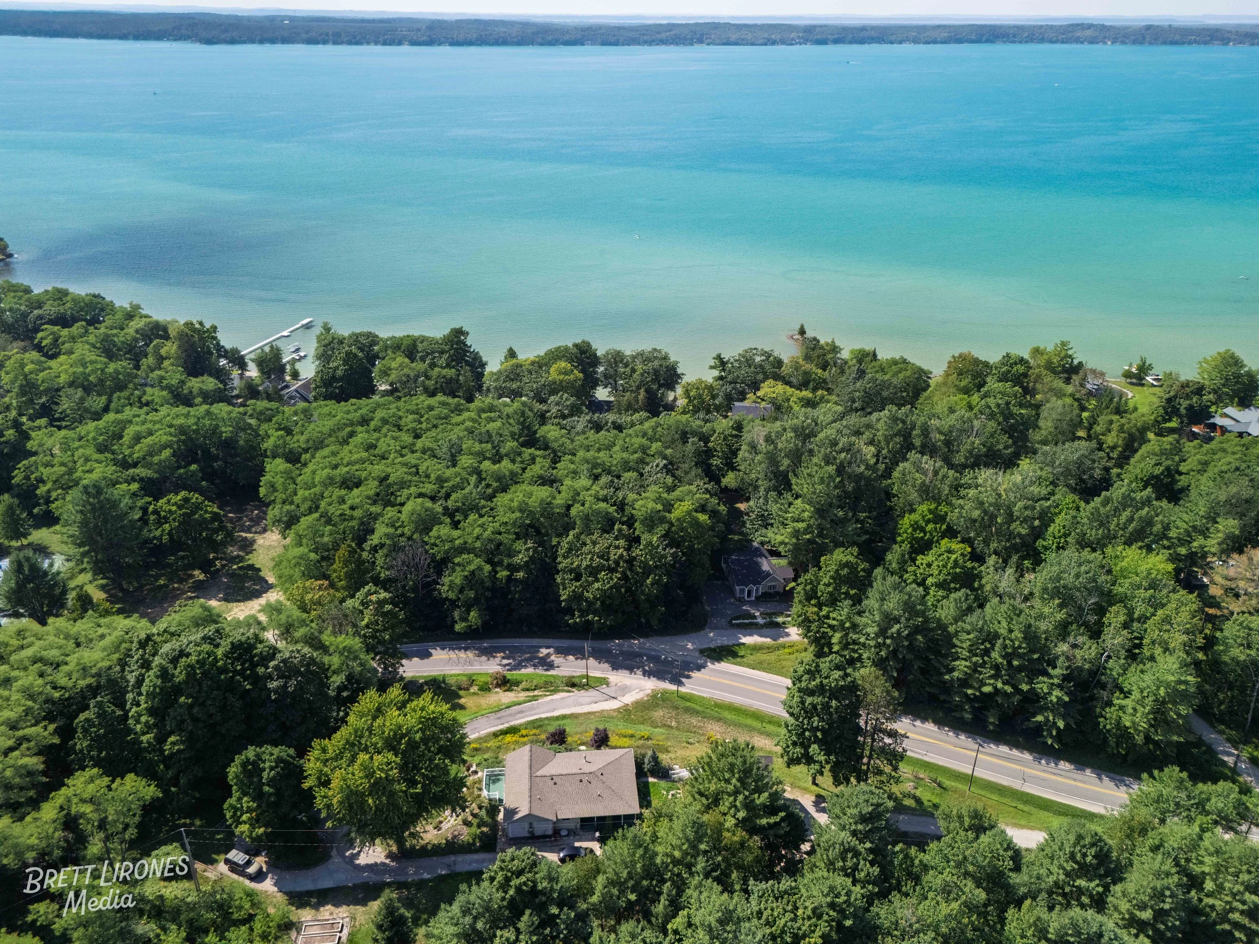 Aerial view of a lakeside scene with a large blue lake, a dock, lush green trees, and some houses along the shoreline.