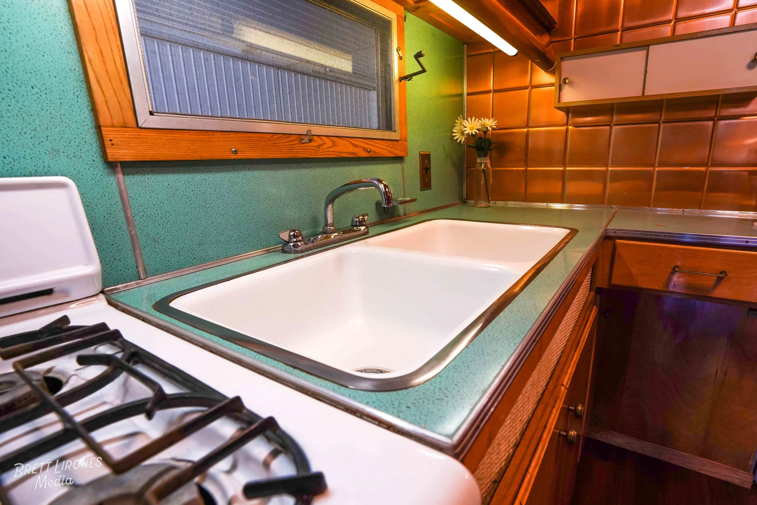 Vintage kitchen with green speckled countertop, white sink, and brown tiled walls, featuring wooden cabinetry and a small window with a wooden frame.