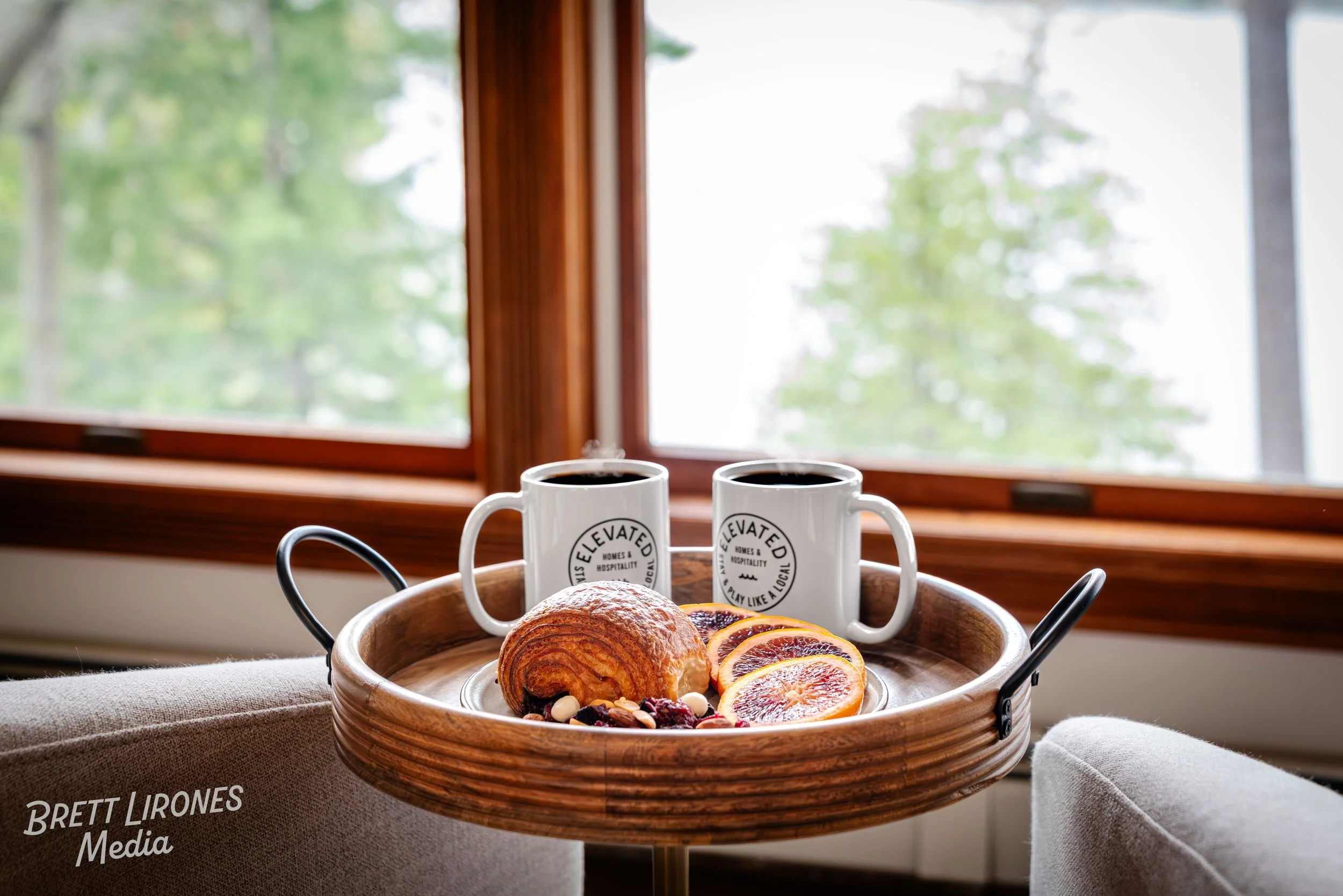 A breakfast tray with croissants, orange slices, fruit, and nuts placed on a table near a window with green trees outside, two coffee mugs with logo, cozy armchair, and natural light.