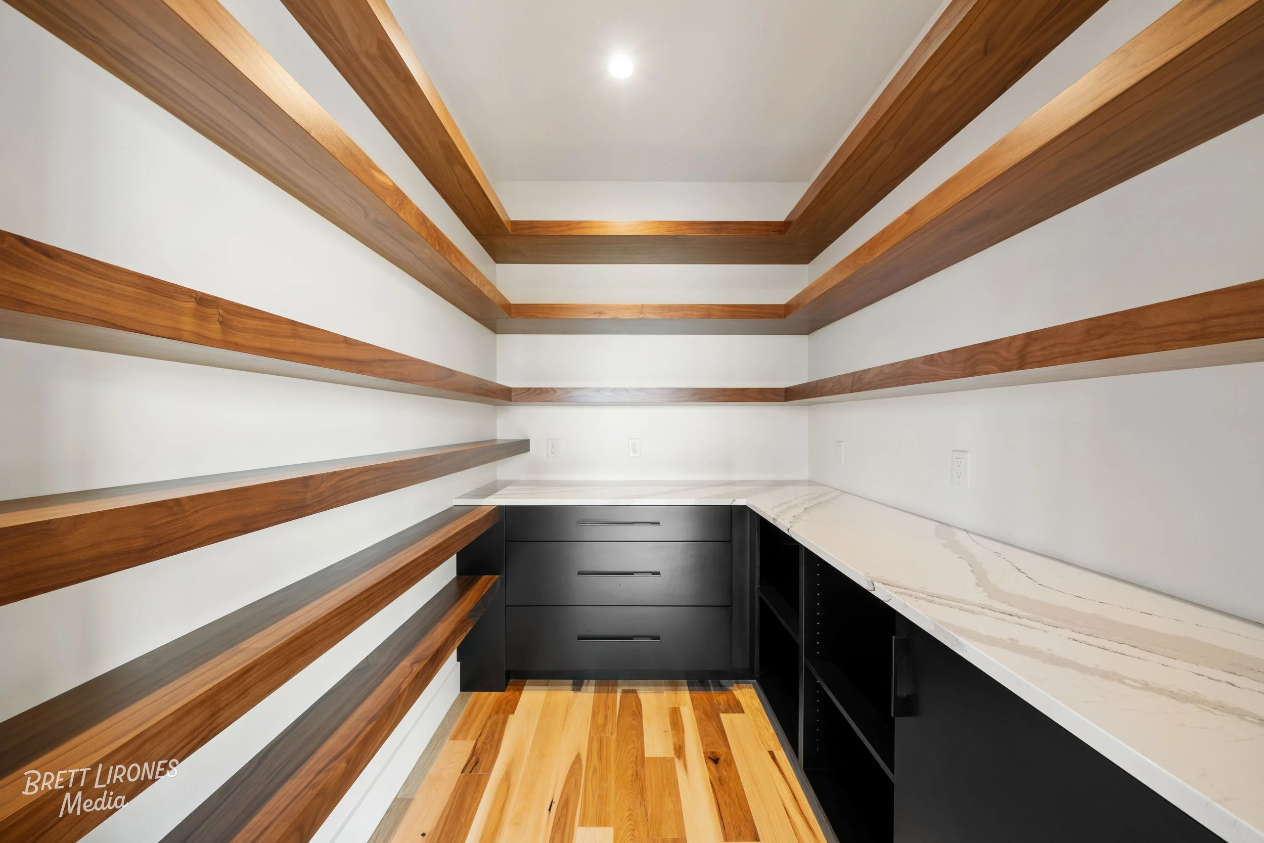 A small walk-in pantry with several wooden shelves on the walls, a black cabinet with drawers, and a marble countertop with a white and beige pattern, with light wood flooring.