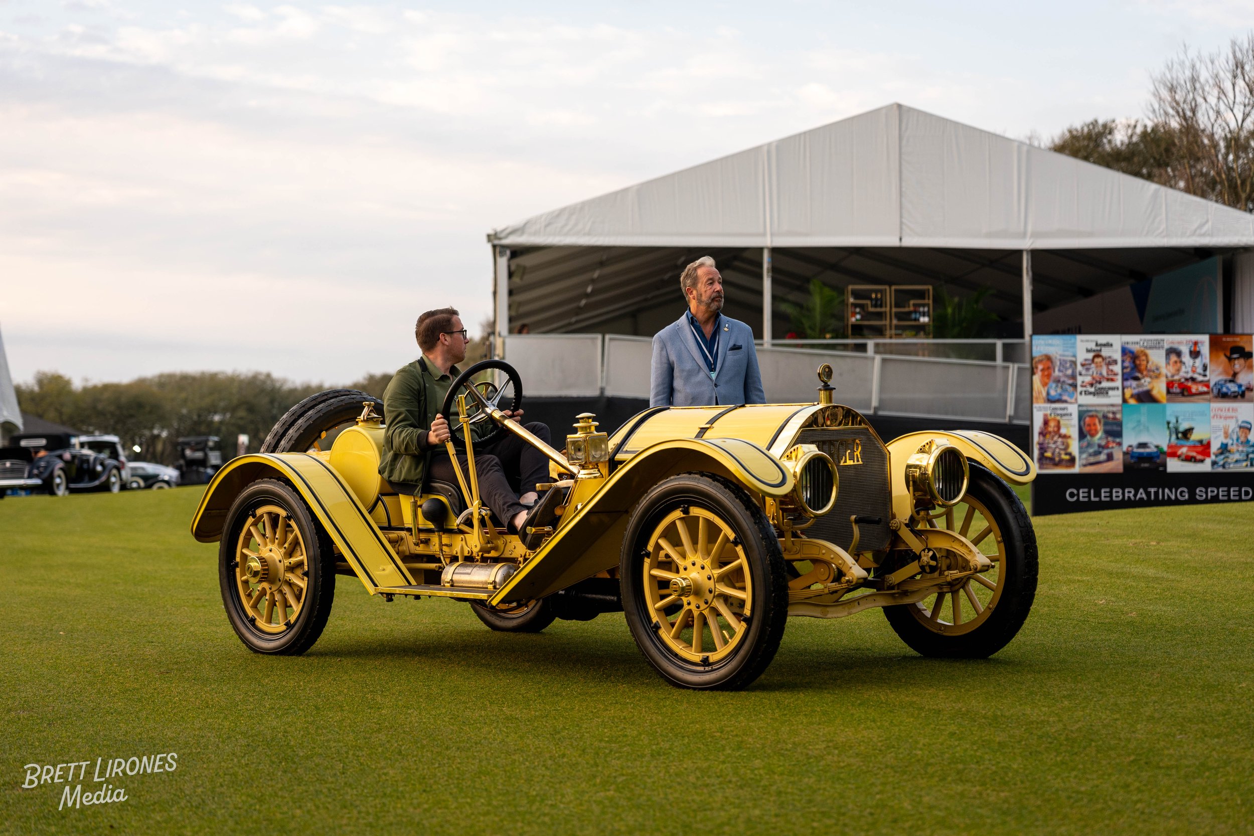 A vintage yellow car with black accents on a grassy field at an outdoor event, with two men near it and a white tent in the background.