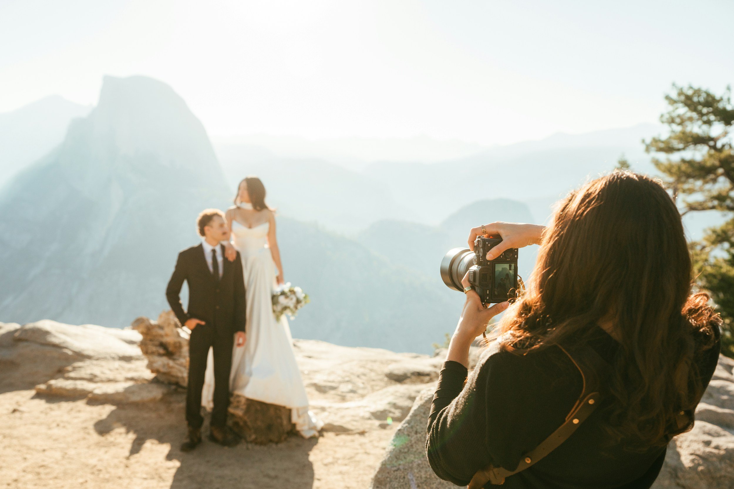 A woman taking a photograph of a couple dressed in wedding attire on a rocky overlook with mountains and a valley in the background.