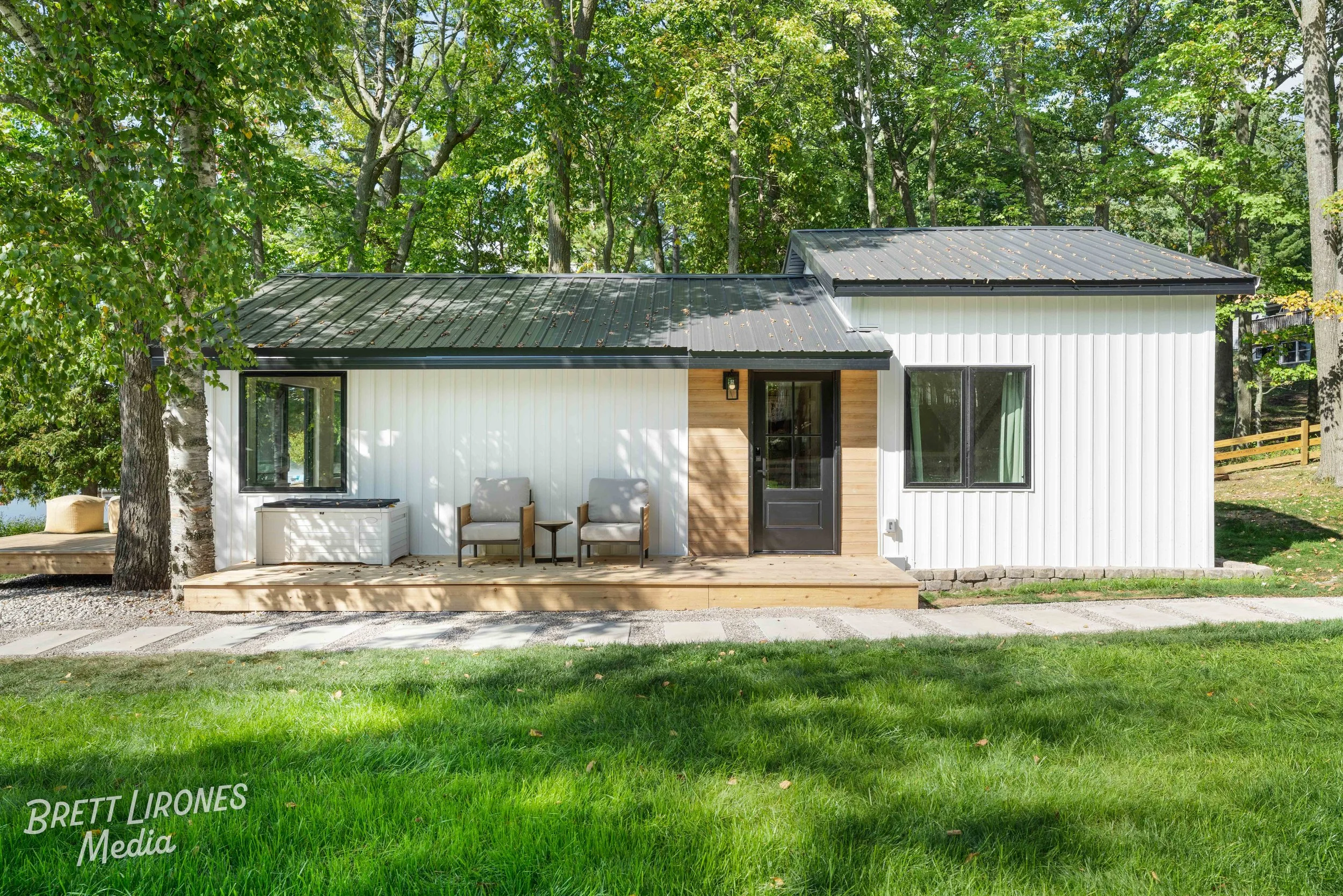 A small modern white house with black door and window frames, set in a green yard with tall trees in the background. It has a wooden porch with two chairs and a side table.