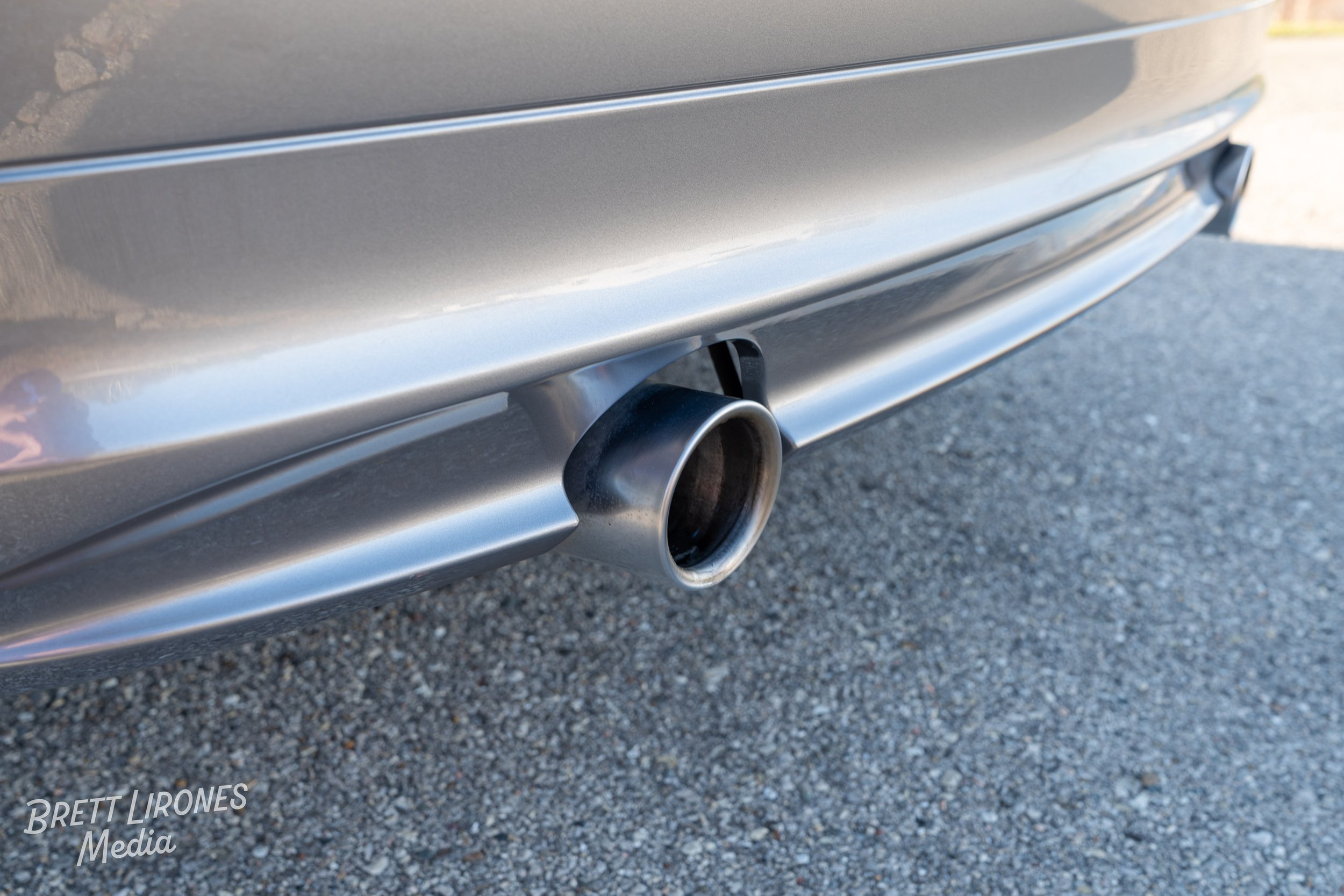 Close-up of a car's exhaust pipe on the rear bumper, with gravel ground underneath.