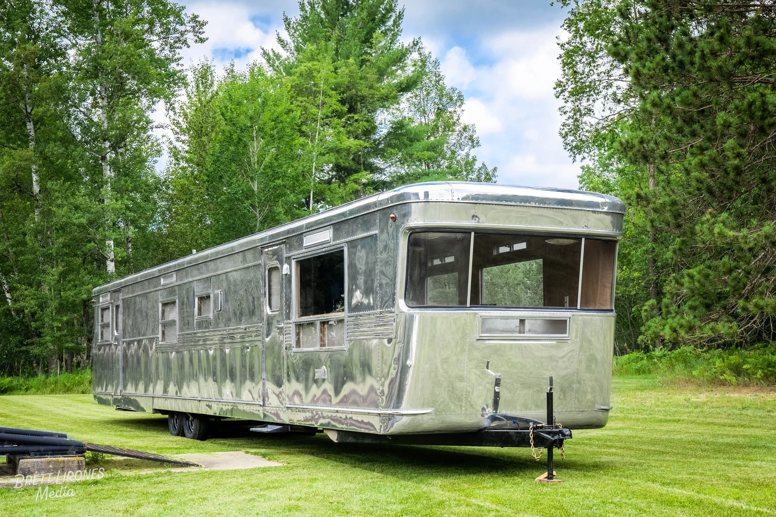 A vintage silver Airstream travel trailer parked on a grassy field with green trees and partly cloudy sky in the background.