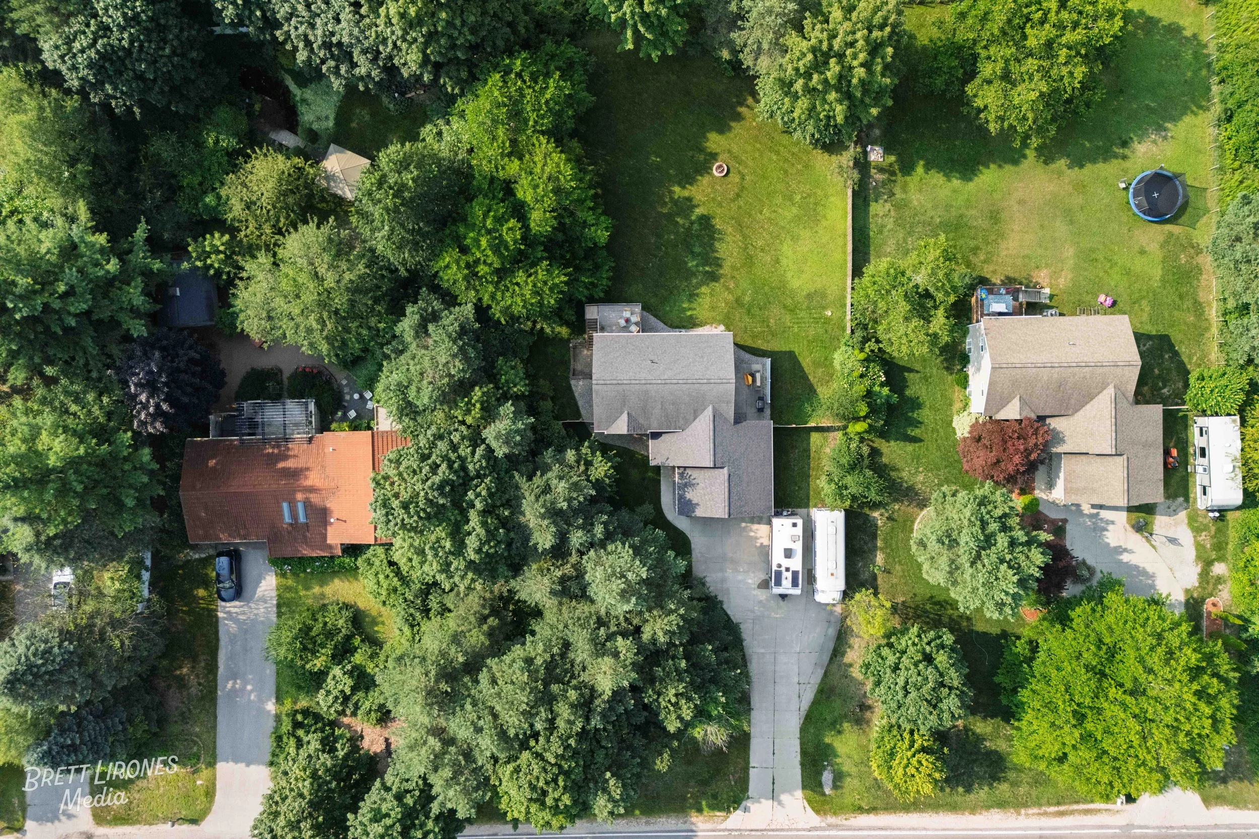 Aerial view of a suburban neighborhood with multiple houses, trees, lawns, and driveways, showing rooftops, a trampoline, and outdoor furniture.