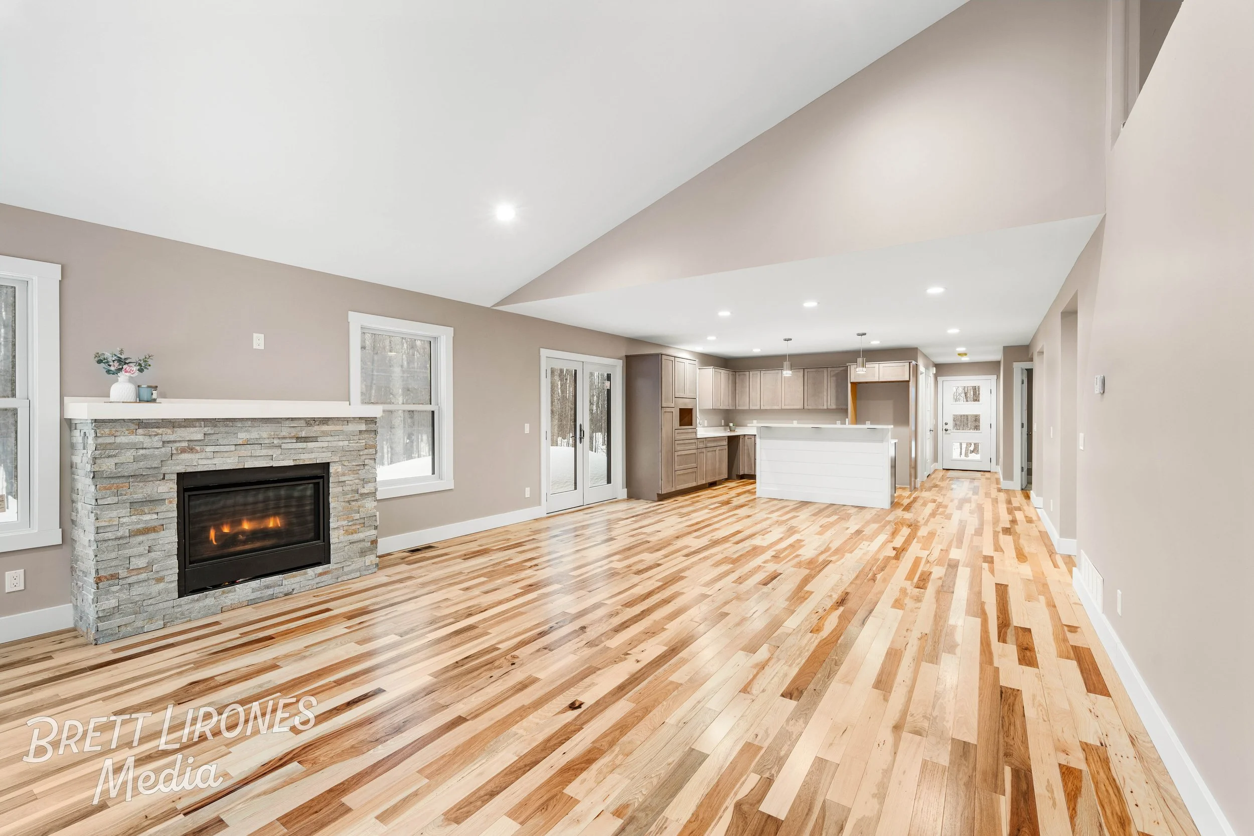 Empty open-plan living space with hardwood floors, a stone fireplace, large windows, and a kitchen with cabinets and a white island.