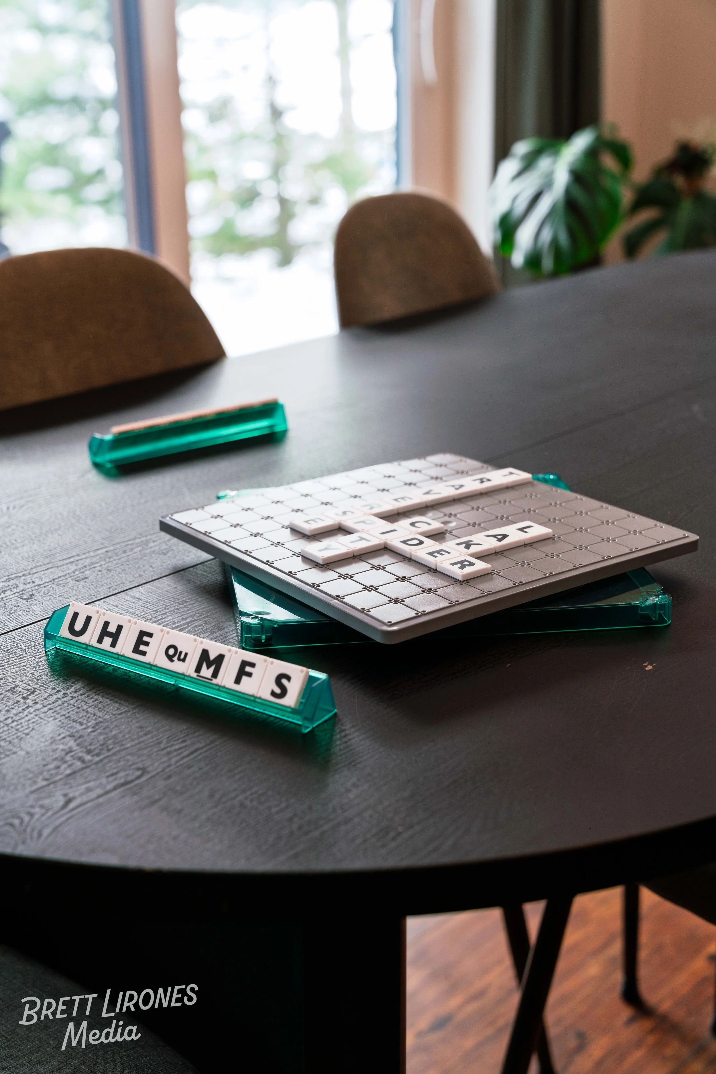 Scrabble tiles on a game board and a tile rack on a dark wooden table near a window with green plants and snow outside.