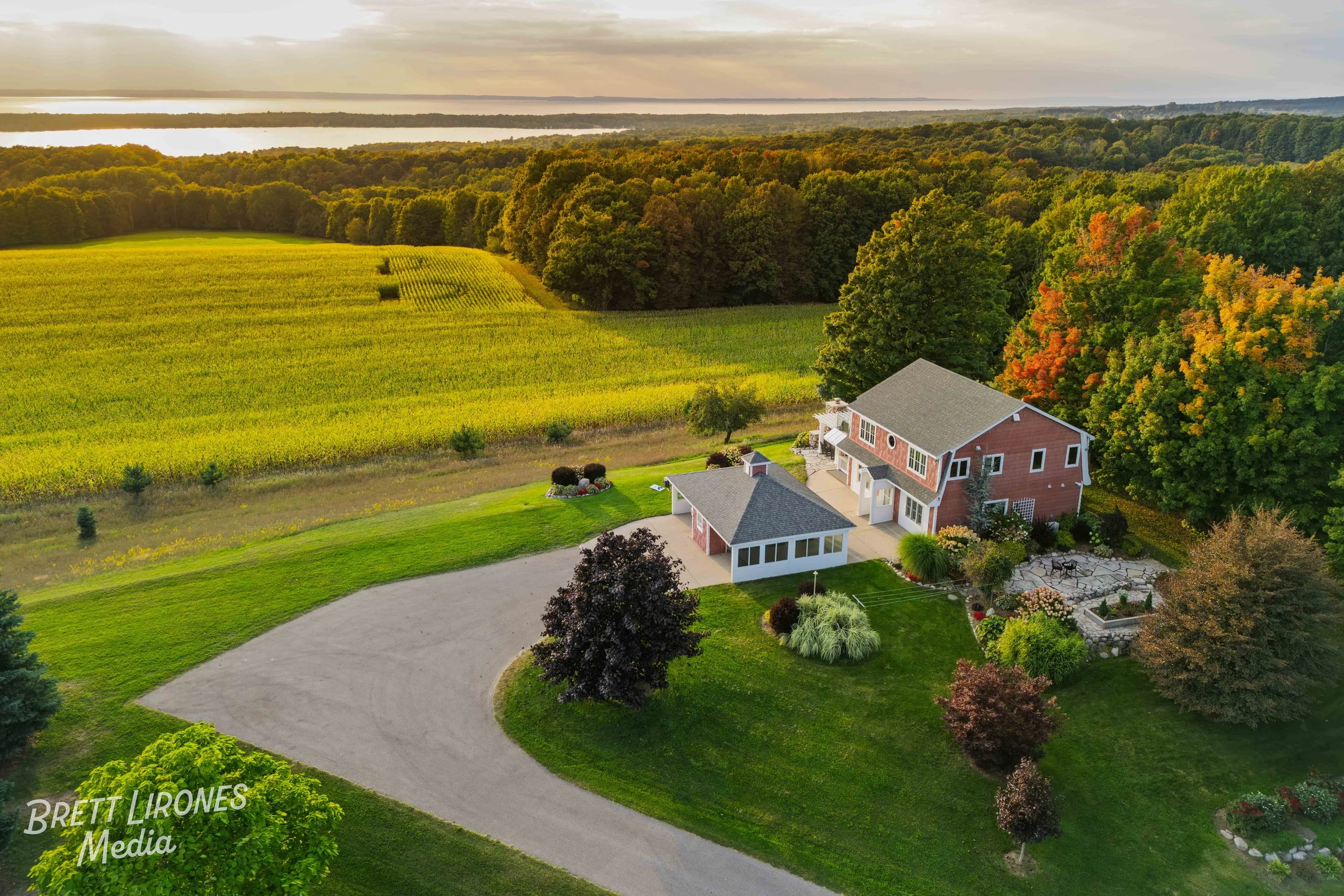 Aerial view of a red house with a gray roof, surrounded by a green yard with trees and a garden, overlooking fields, a lake, and a forest in the distance during sunset.