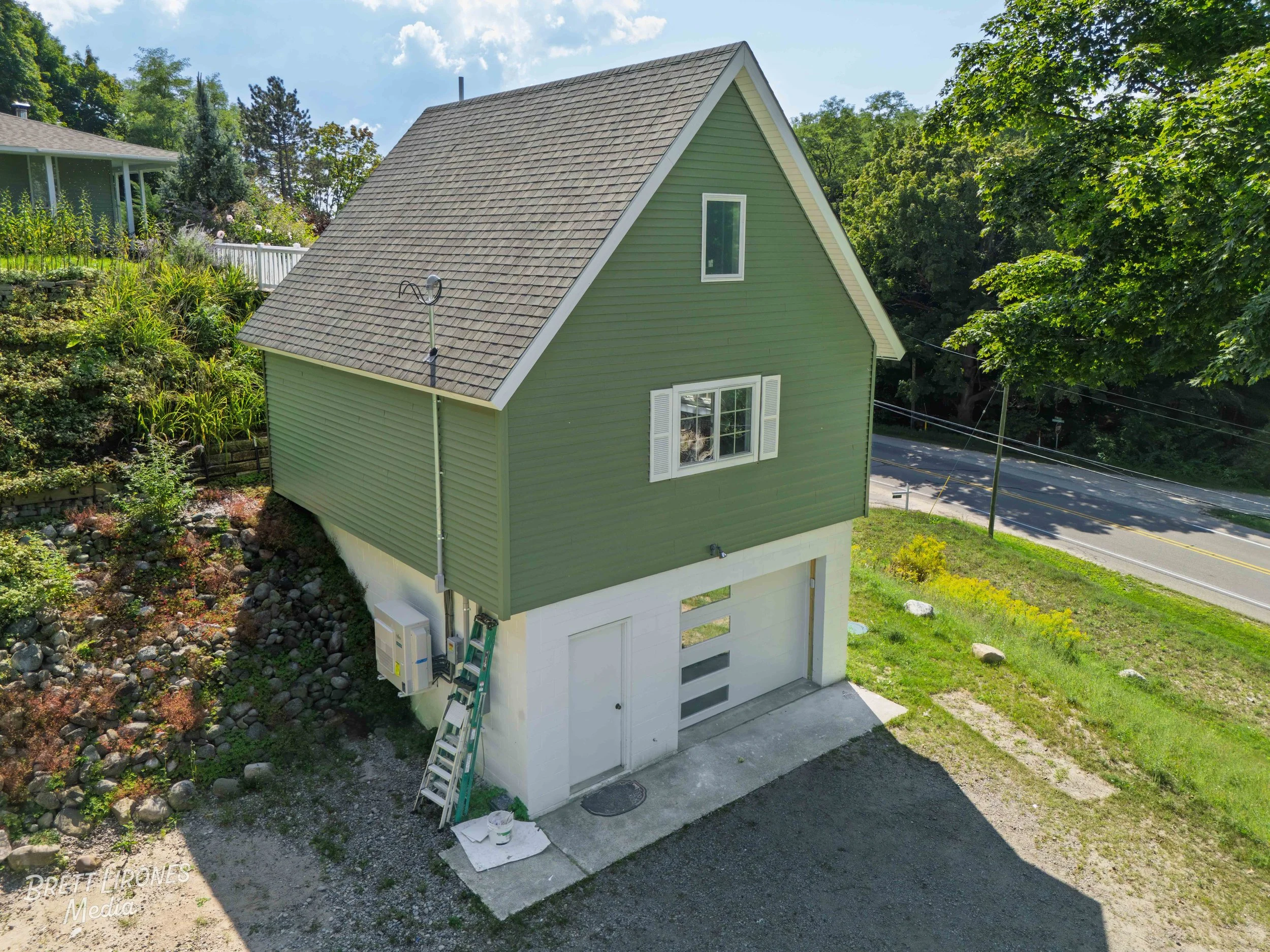 A green, two-story house with a garage on the ground level, located on a sloped lot surrounded by trees and greenery. The house has white trim, a window with shutters, and a ladder leaning against the side of the garage.
