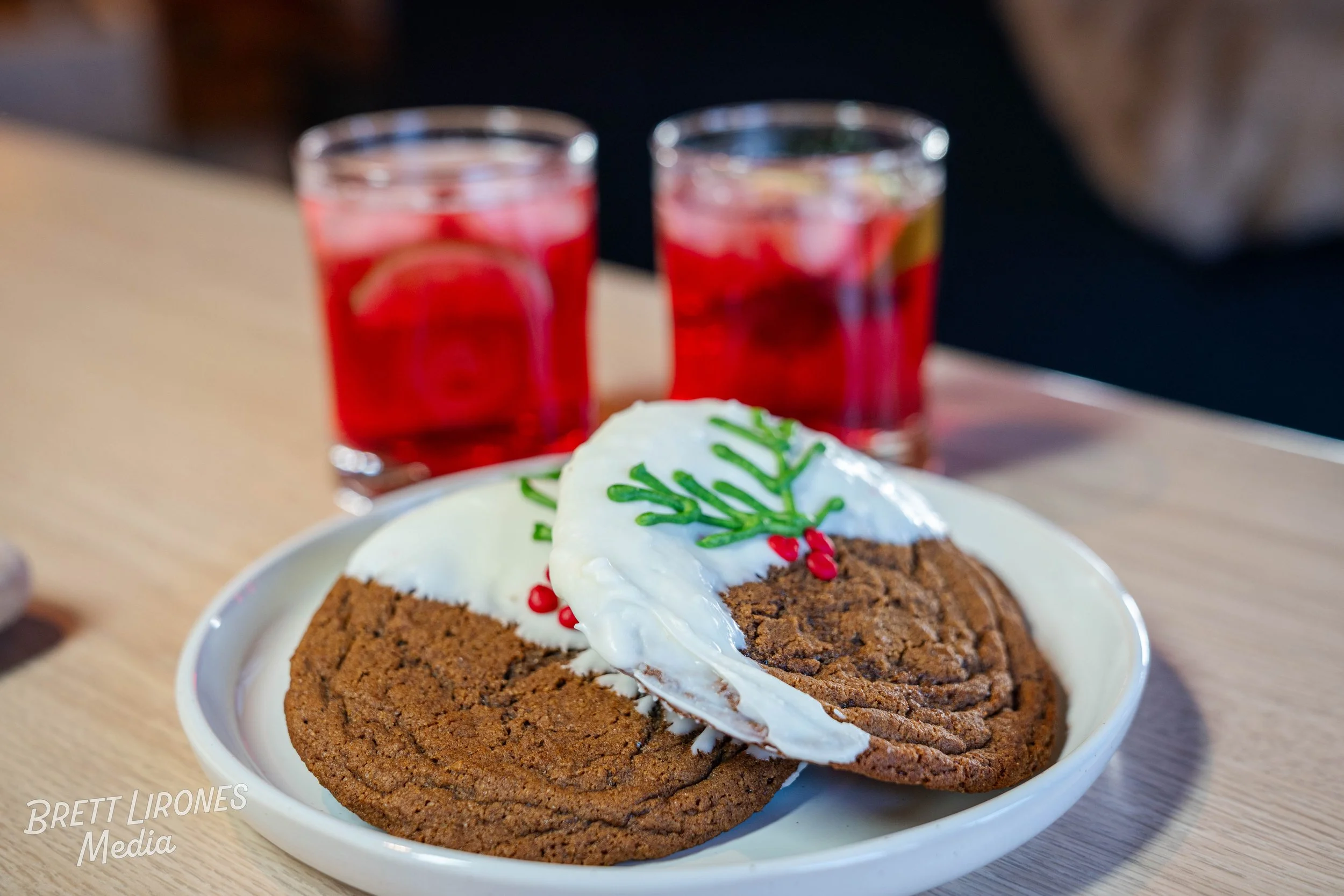 A white plate with two large chocolate cookies topped with whipped cream, decorated with green sprigs and red berries, on a light wood table. In the background, two glasses of a red beverage with lemon slices.