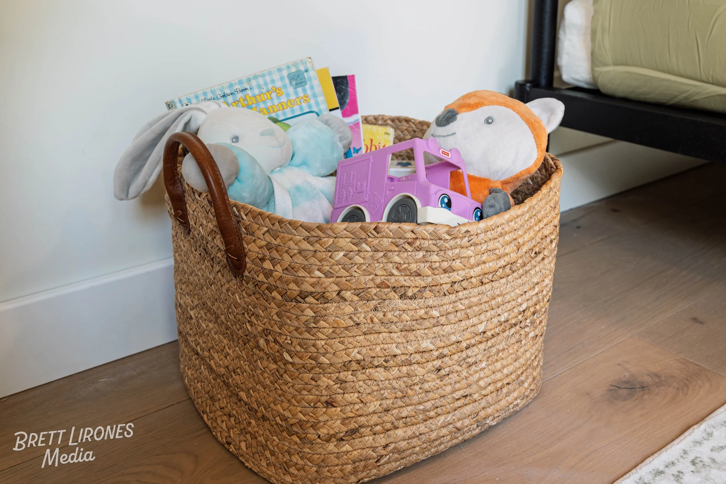 A woven basket containing stuffed animals, a purple toy car, and books placed on a hardwood floor