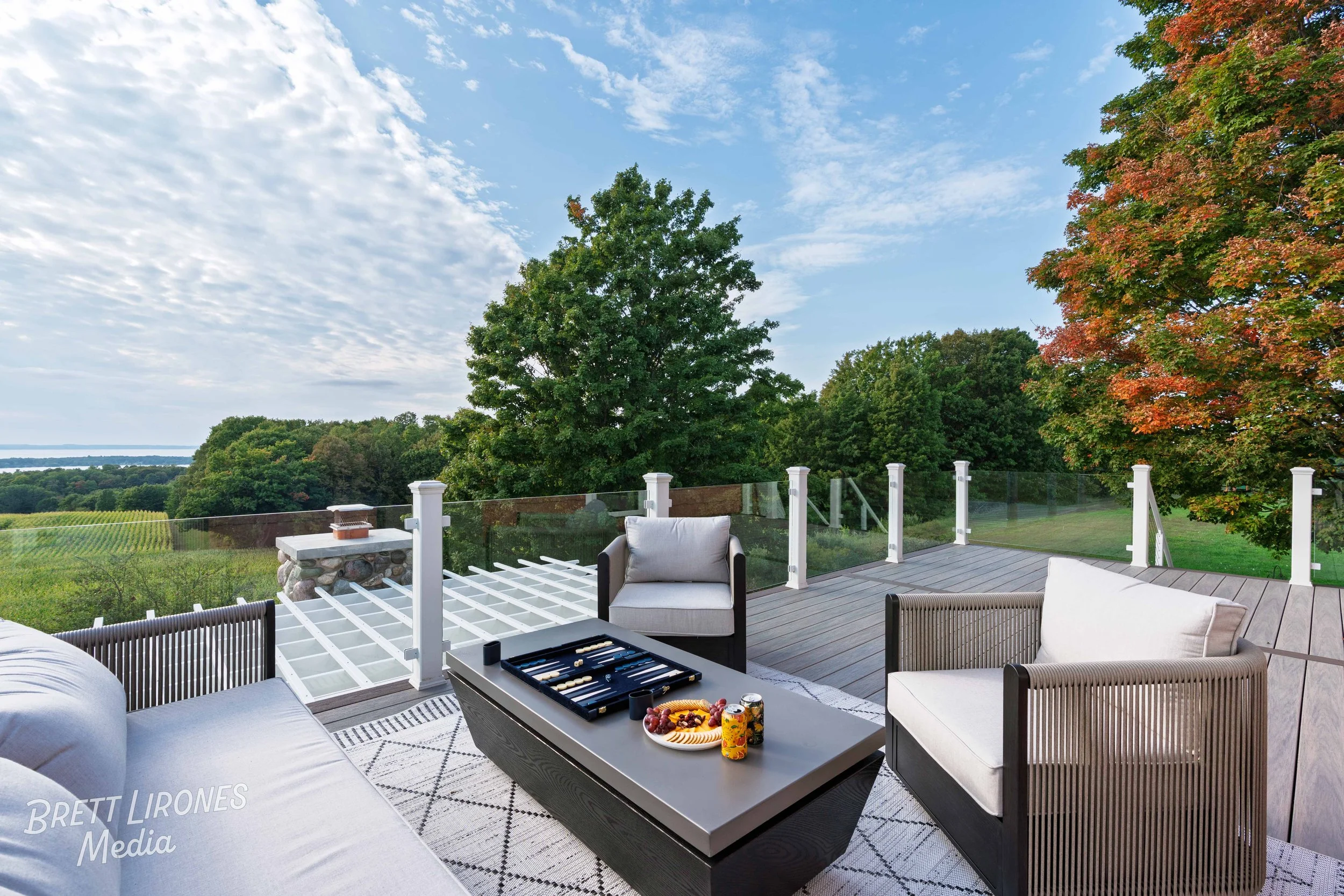 Outdoor patio with white cushioned chairs and a table with a backgammon game and snacks, overlooking green trees and a blue sky with clouds.