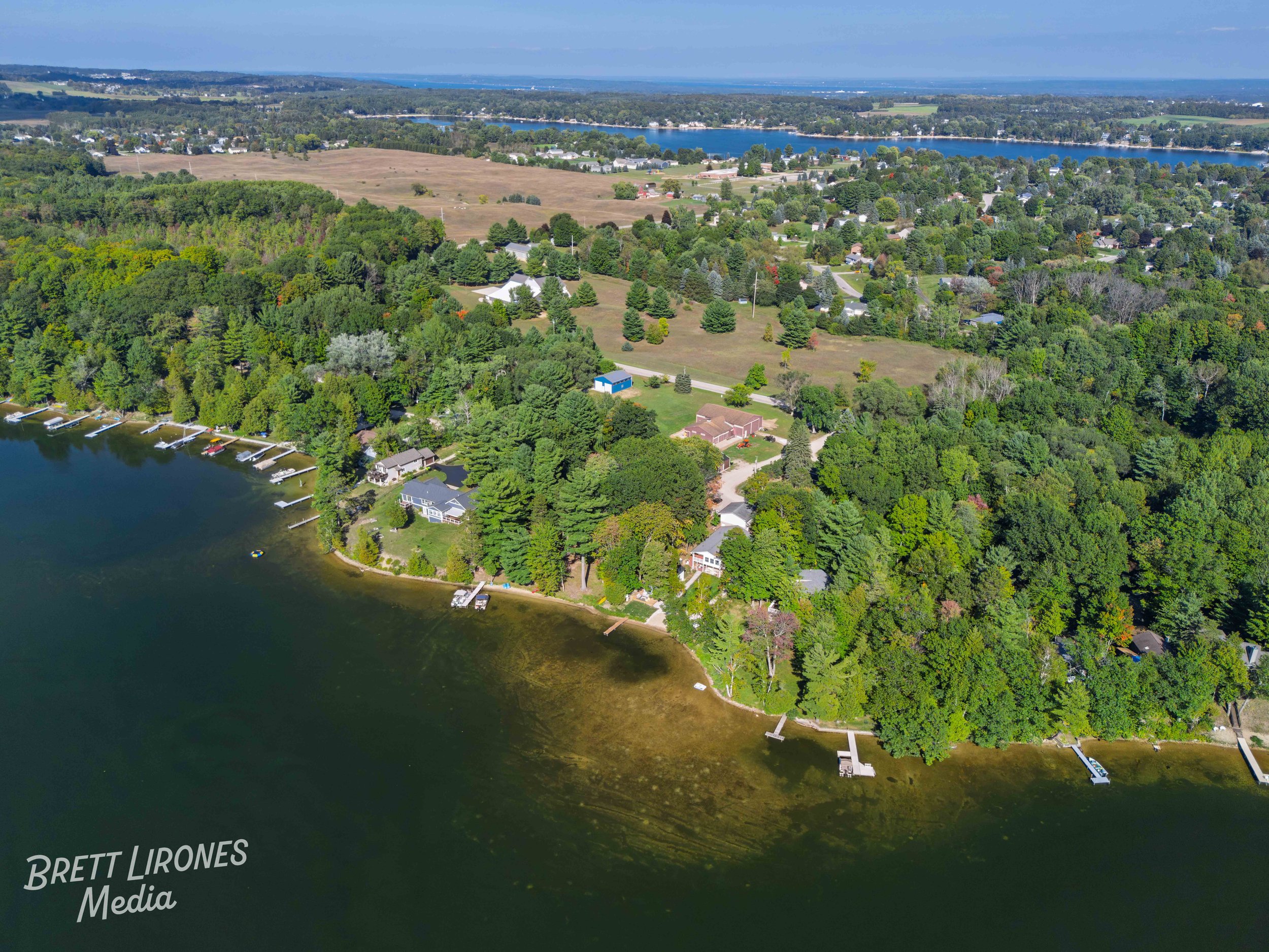 Aerial view of a lakeside residential area surrounded by green trees, with houses along the shoreline, dock structures, and boats, extending towards a larger body of water in the distance.