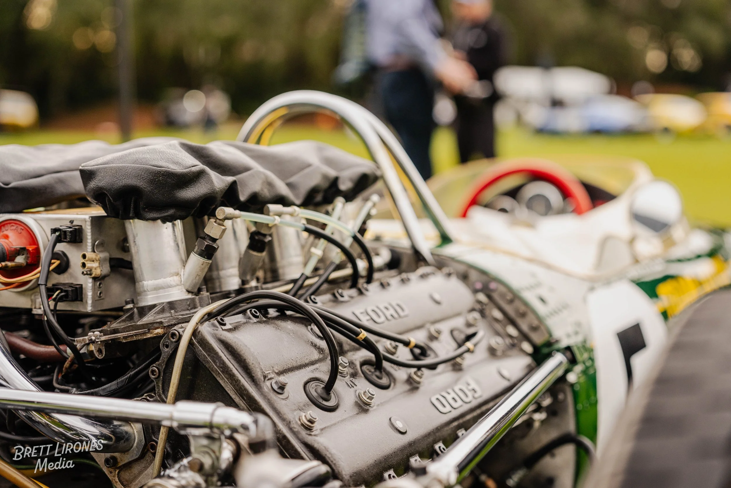 Close-up of a vintage race car engine with visible spark plug wires and mechanical components, with a blurred background of people and other cars.