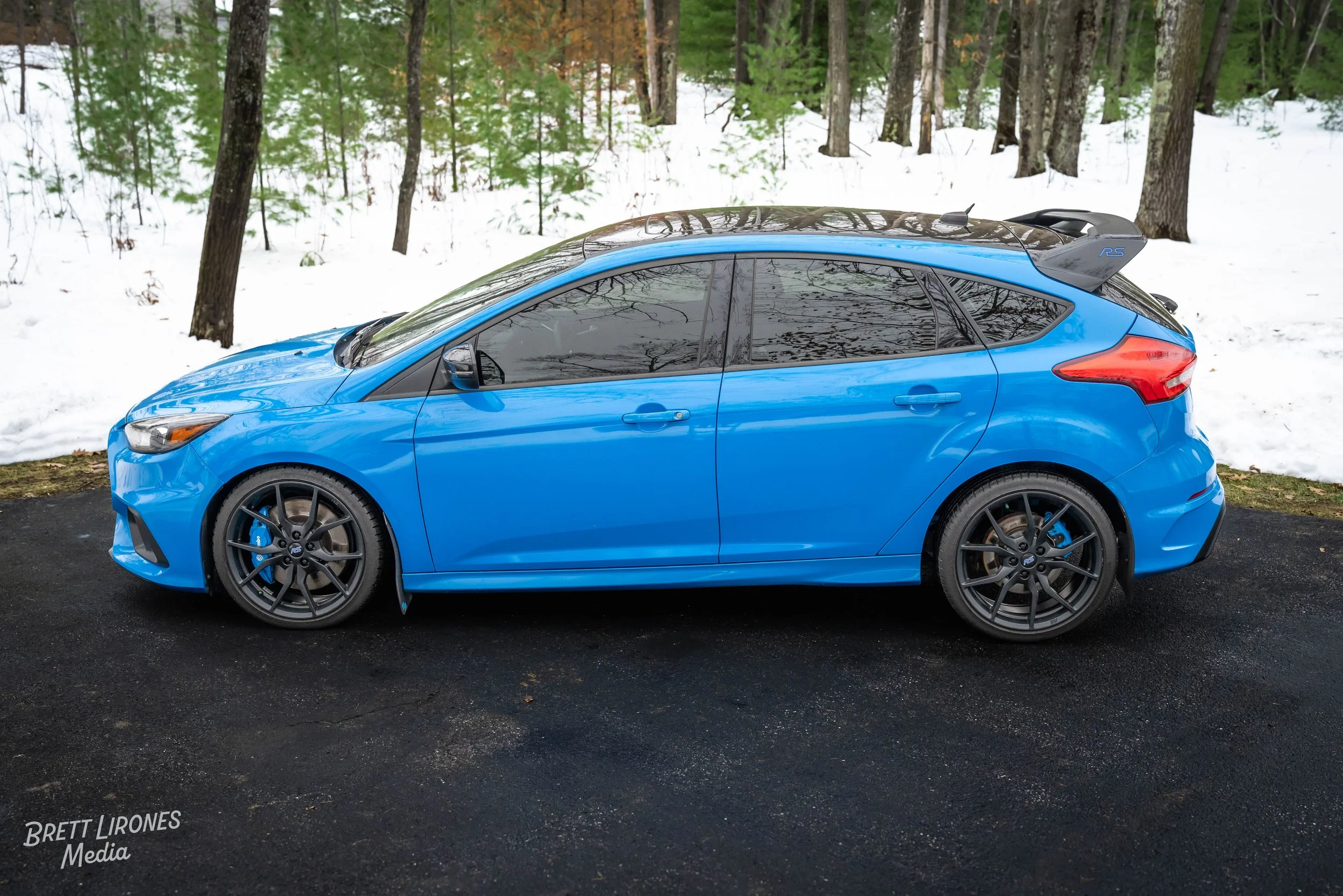 A blue hatchback car parked on a paved surface with snow and forest trees in the background.