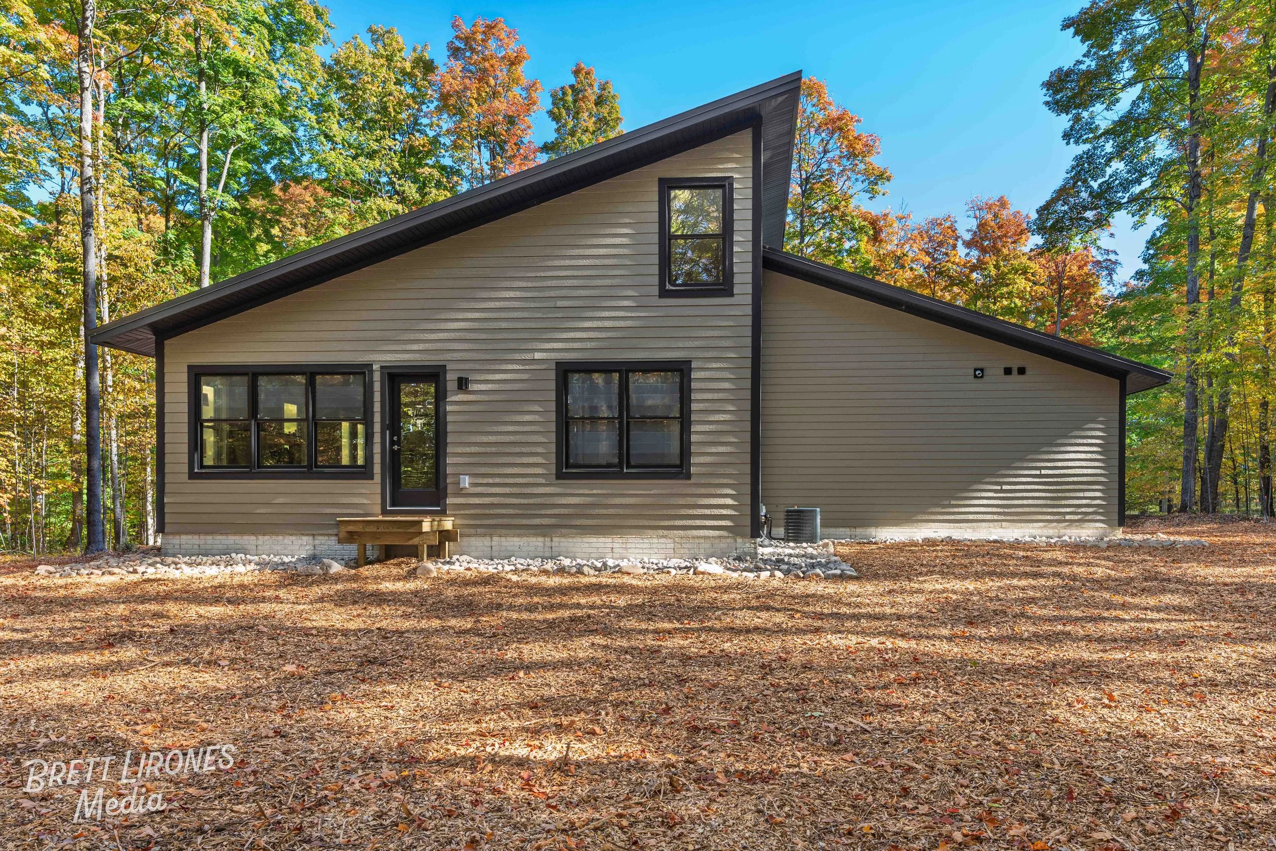 A modern house with beige siding and black window frames, situated in a wooded area with colorful autumn trees, a blue sky, and a dirt yard in the foreground.