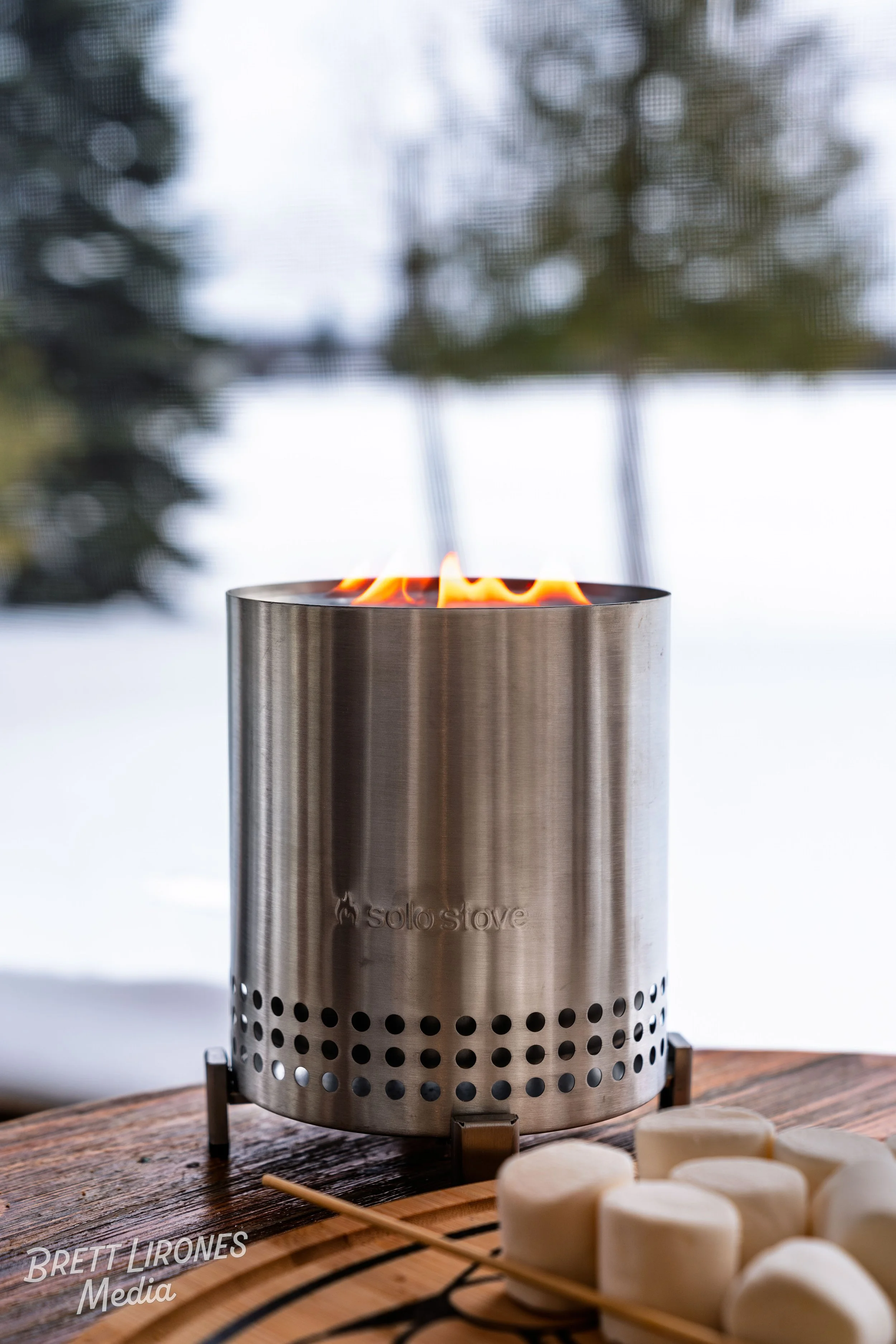 A stainless steel Solo Stove with visible flames on a wooden table, nearby marshmallows on a skewer, with a blurred outdoor background.