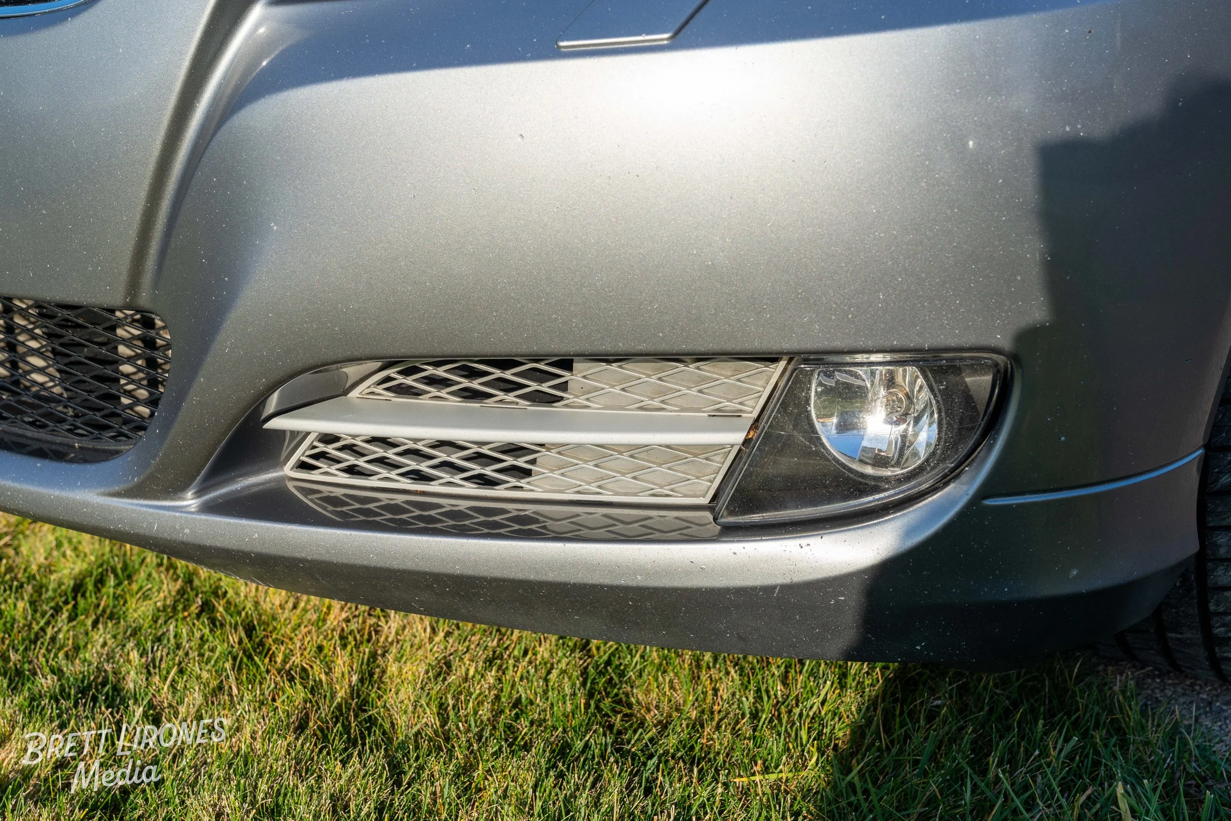 Close-up of the front right corner of a silver car showing a headlight, grille, and part of the tire on a grassy surface.