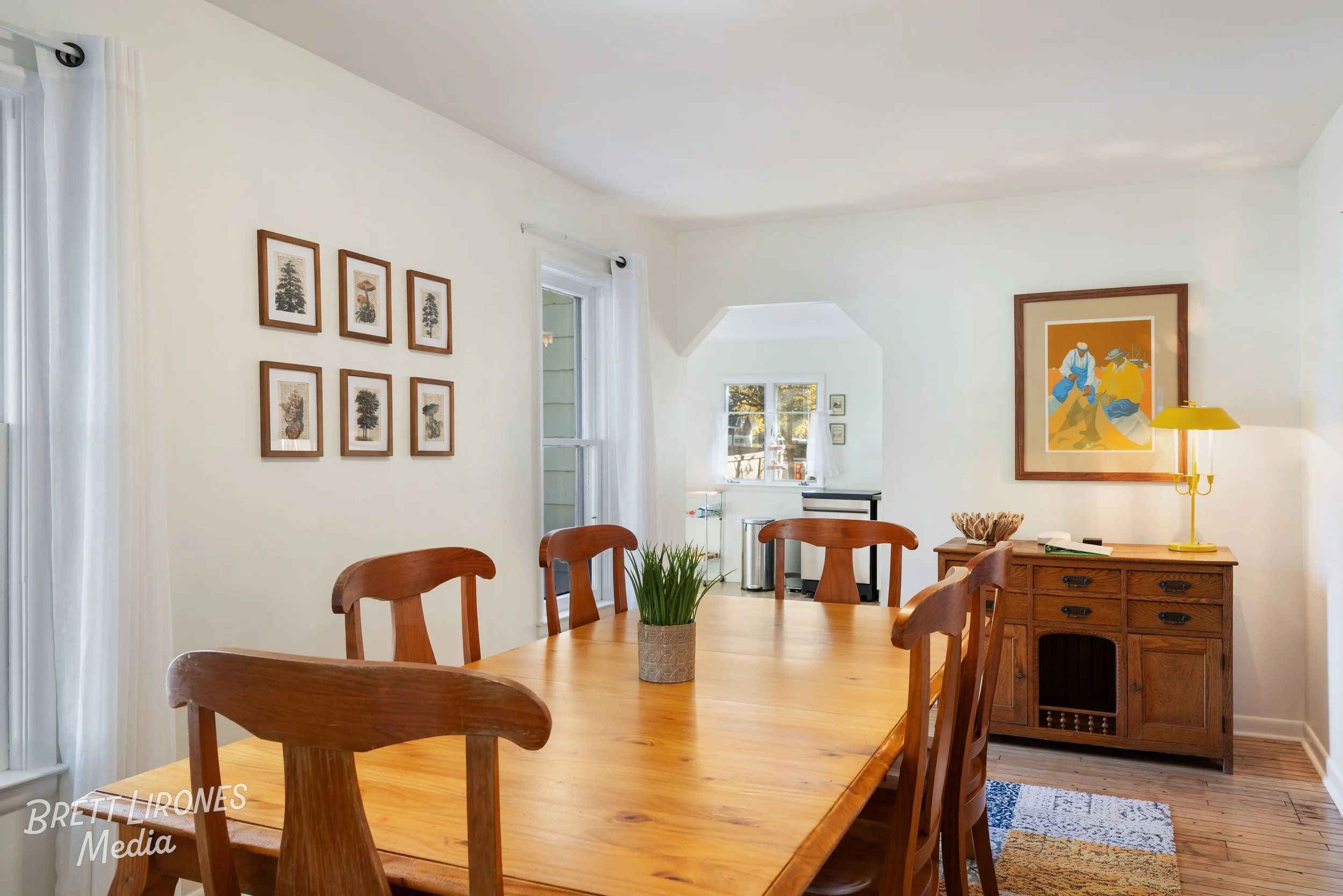 Wooden dining table with six chairs, beige walls with framed art, a wooden sideboard with a yellow lamp, and a small window in a bright, cozy dining room.