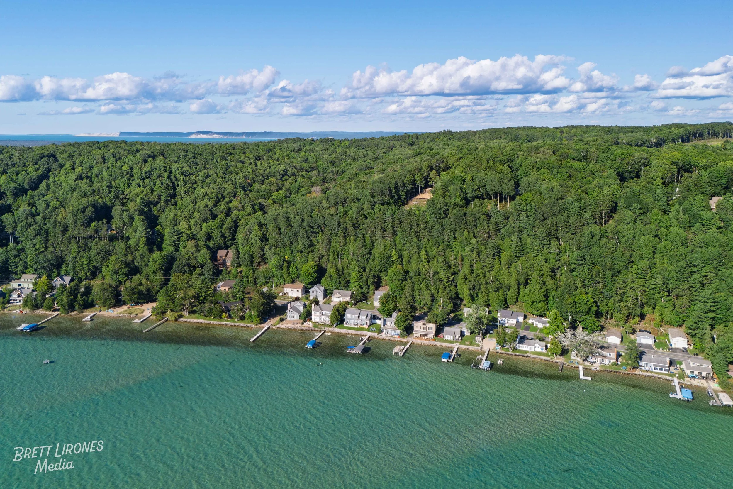 Aerial view of a coastal neighborhood with houses along the shoreline, green forested hills in the background, and a partly cloudy sky.