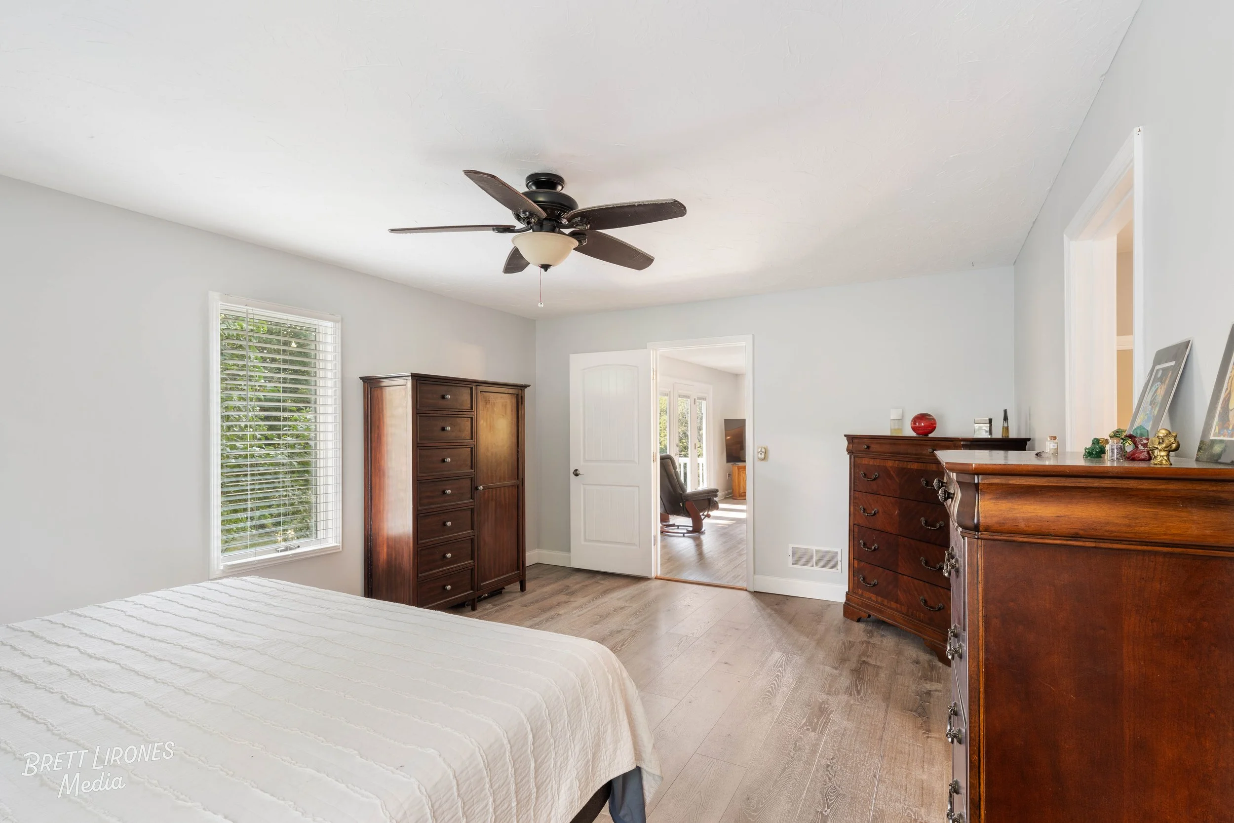 Bedroom with a bed, wooden dresser, chest of drawers, window with blinds, and a ceiling fan, with an adjacent room visible through an open door.