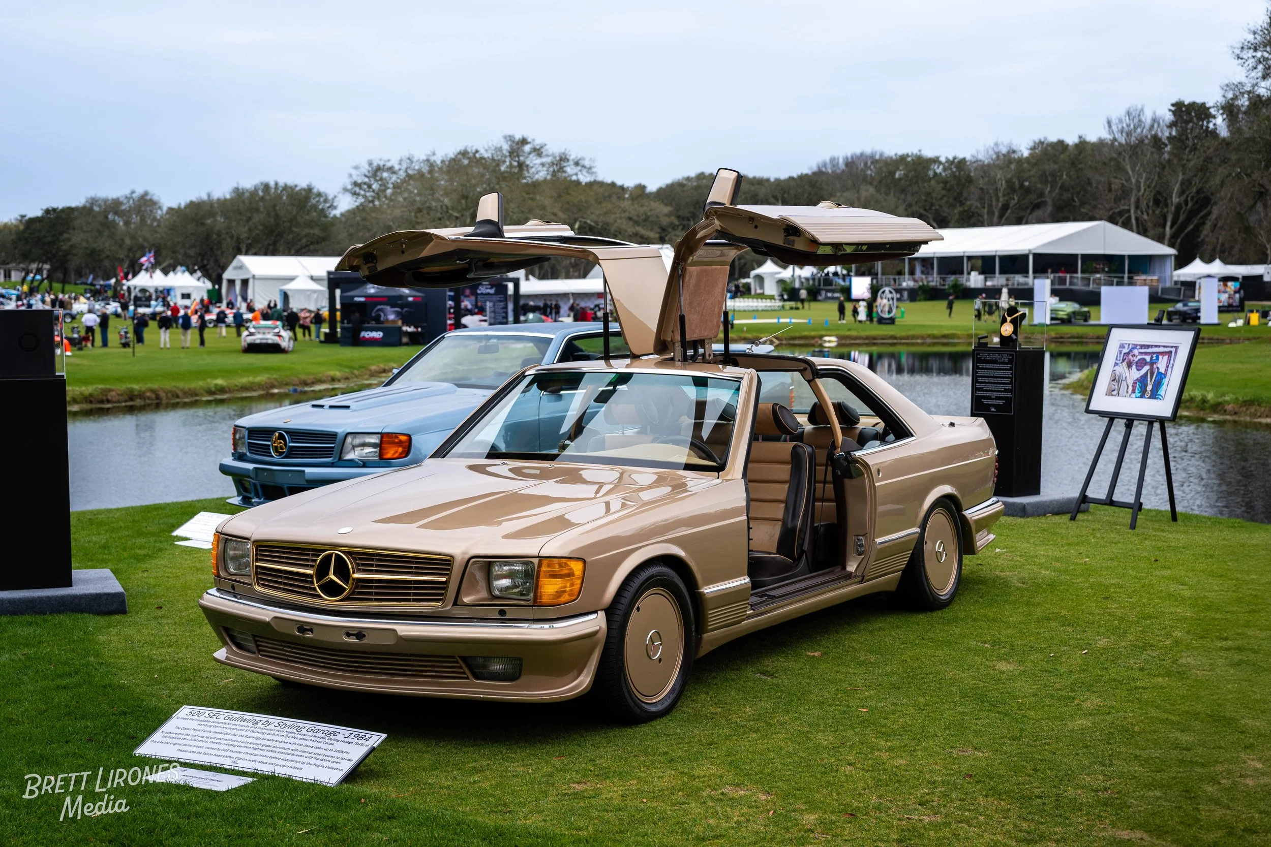 A beige classic Mercedes-Benz coupe with gullwing doors open on display at an outdoor car show, with a blue car and tents in the background near a pond.
