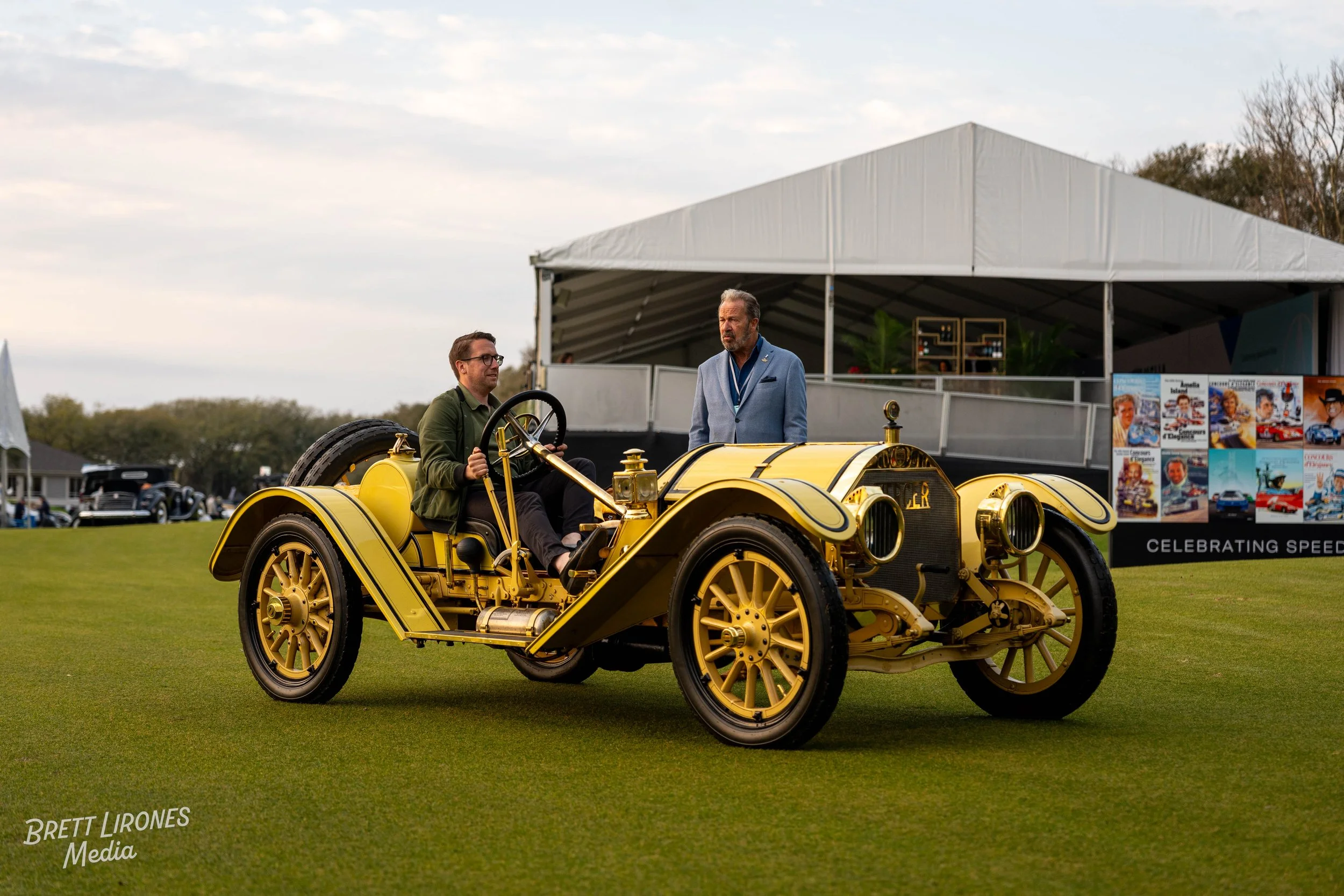 A vintage yellow and black racing car with two men beside it, one seated and one standing, on a grassy field at an outdoor event with a large white tent in the background.