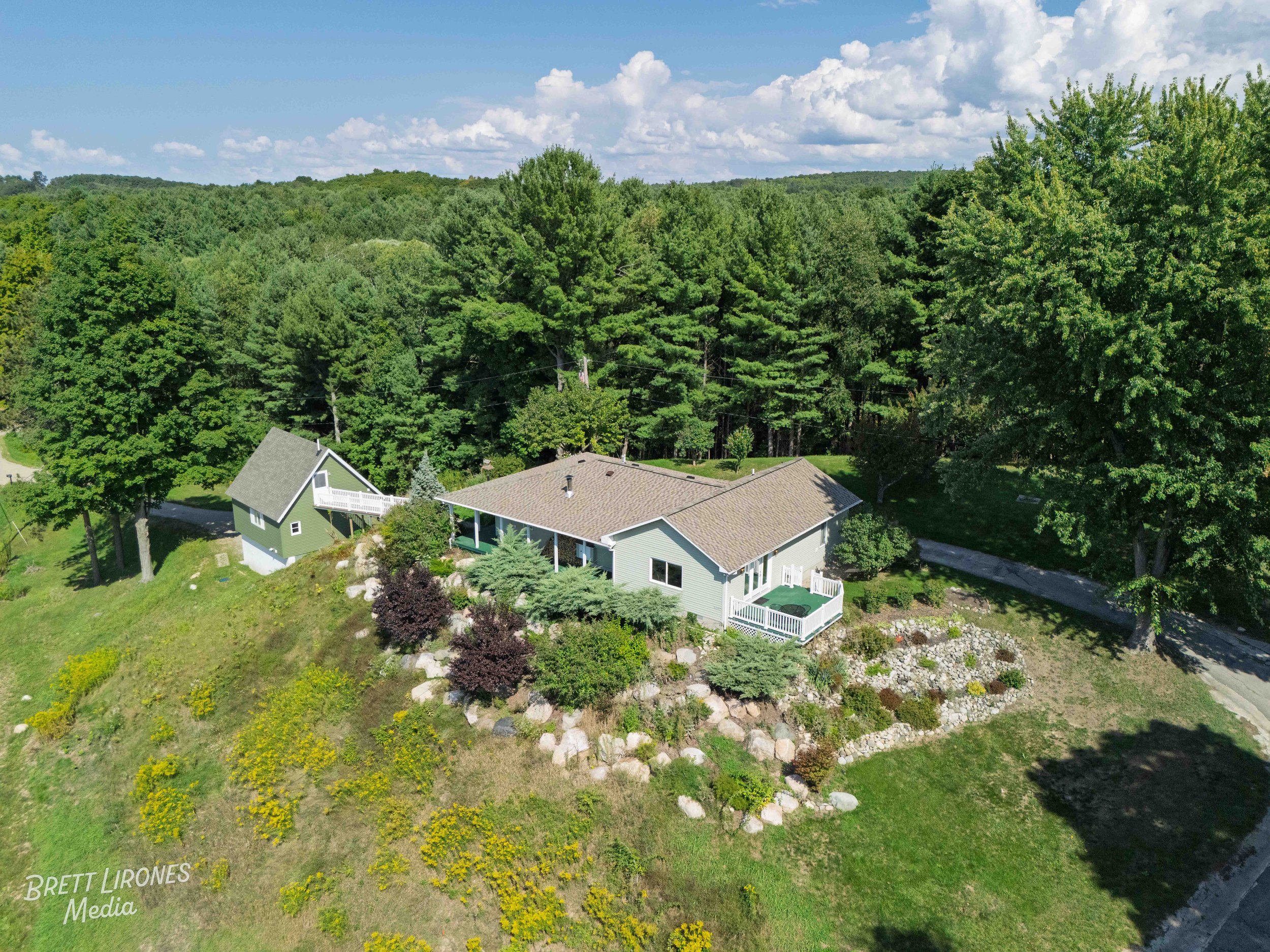 Aerial view of a single-family house surrounded by lush green trees and colorful bushes in a rural setting with a dirt road nearby.