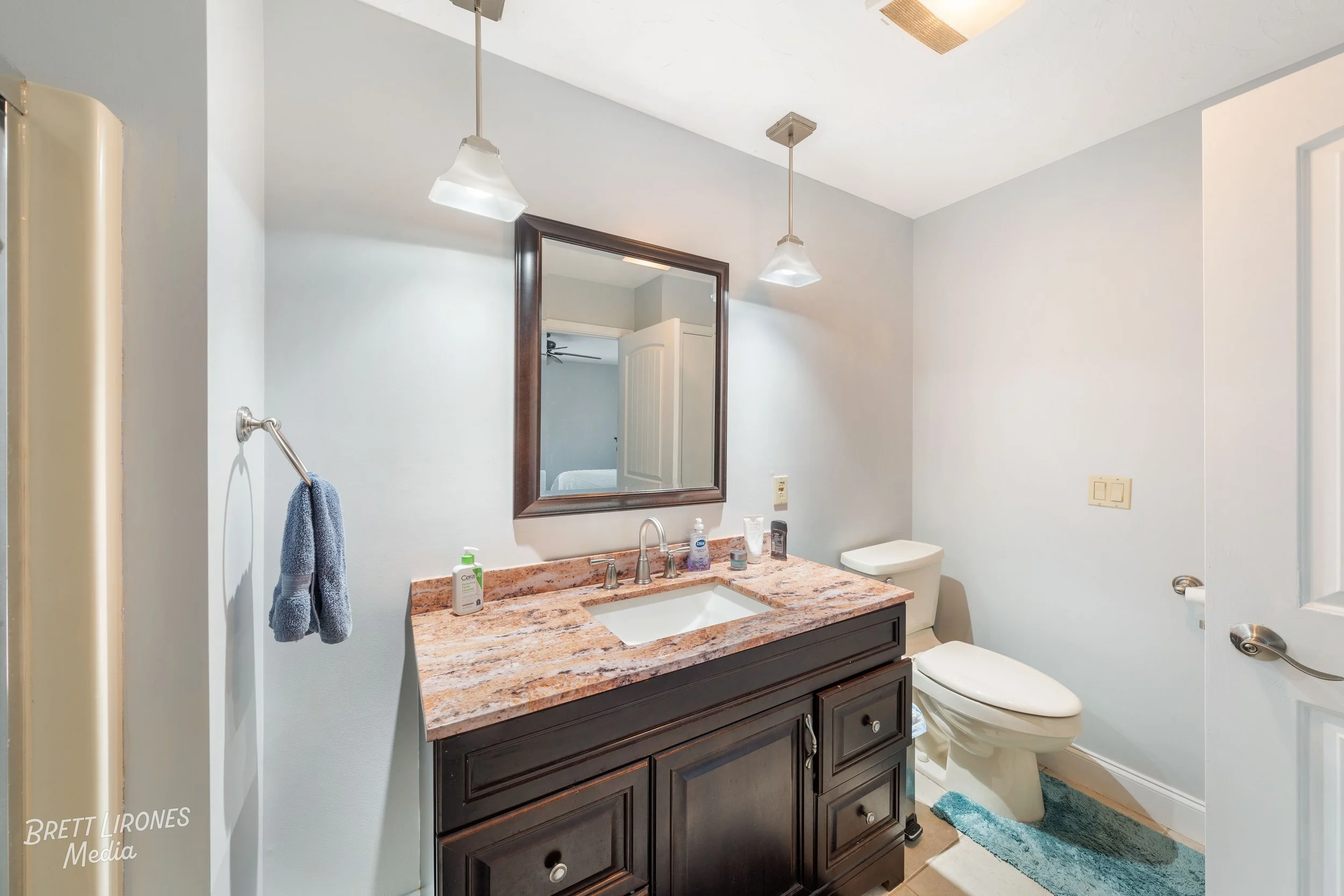 A bathroom with a dark wood vanity, a granite countertop, a mirror, two hanging lights, a towel ring with a blue towel, and a toilet with a blue rug.