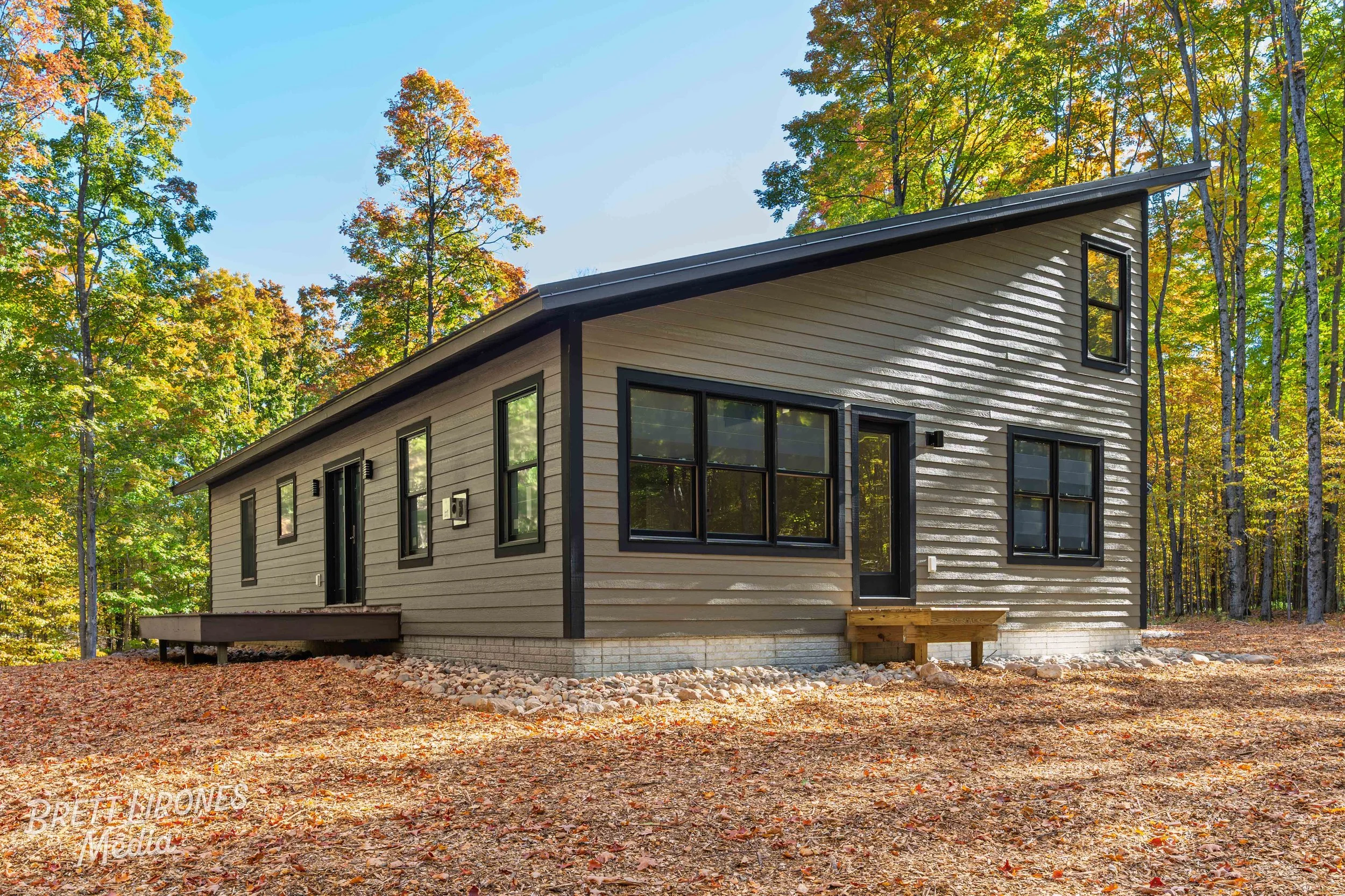 A modern, beige house with black window and door trims situated in a wooded area with autumn foliage, surrounded by fallen leaves and trees.