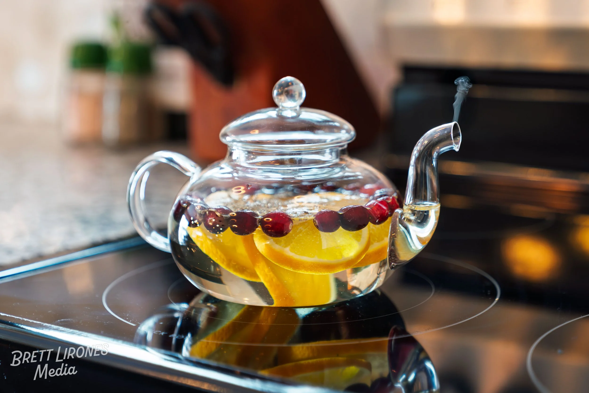 Glass teapot with sliced lemons and cranberries inside, placed on a black stovetop.