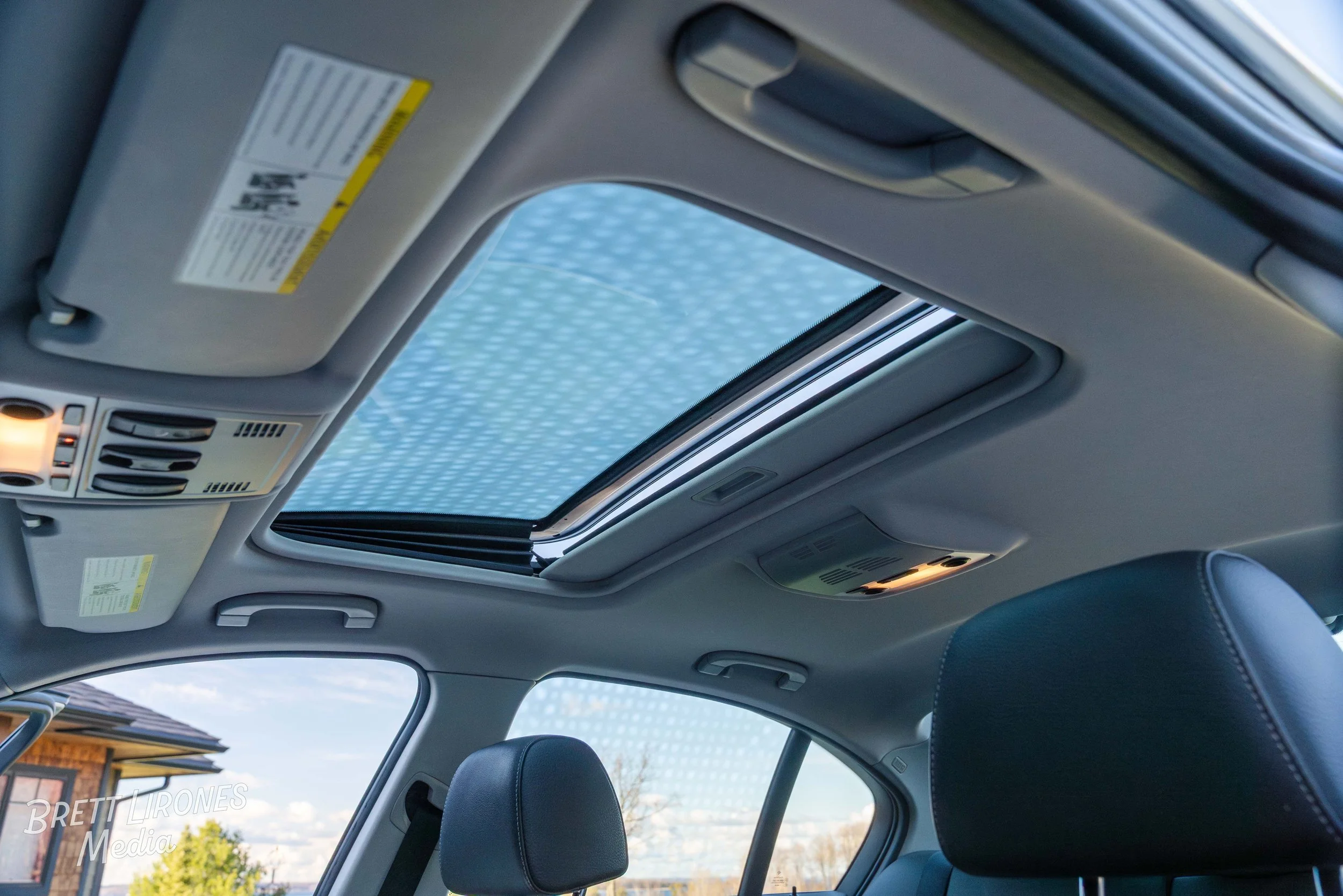 The interior ceiling of a vehicle showing a sunroof, overhead lights, and control panels, with the sky and part of a building visible through the sunroof.