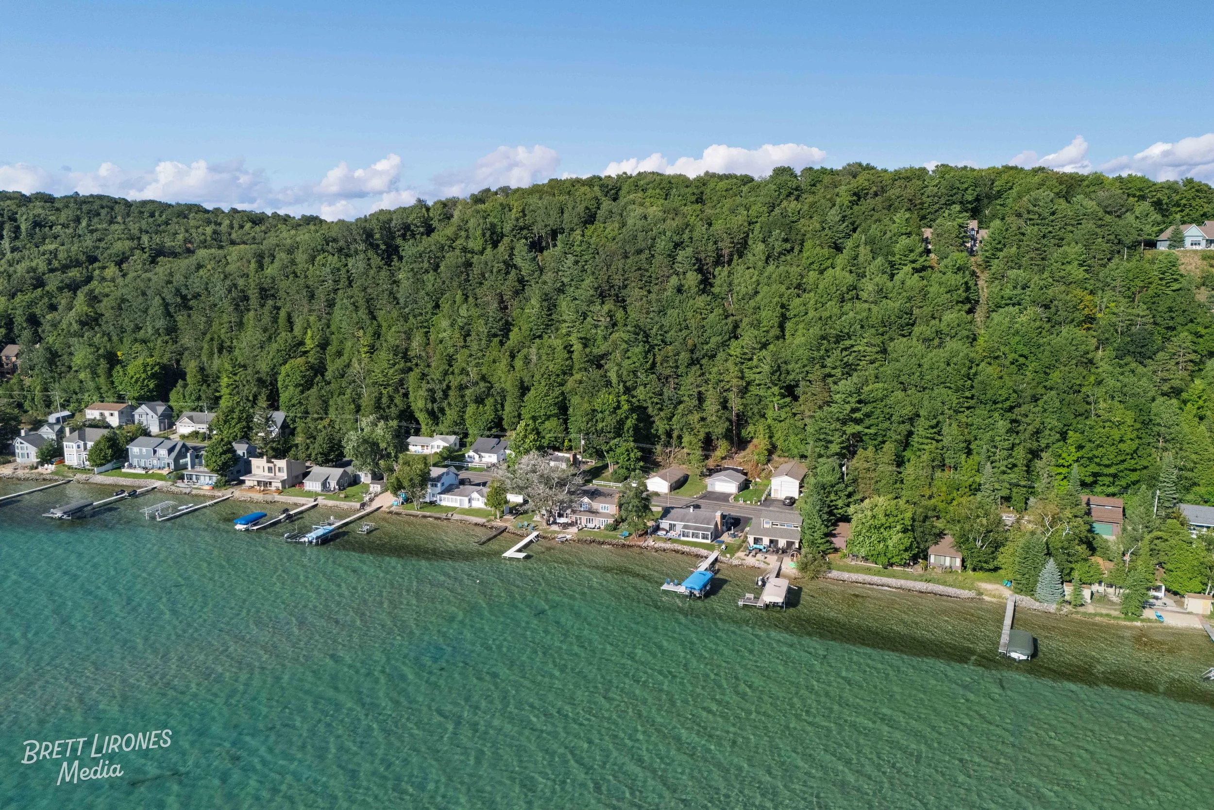 Aerial view of a lakeside community with houses and boat docks along the shoreline, backed by a wooded hillside under a partly cloudy sky.