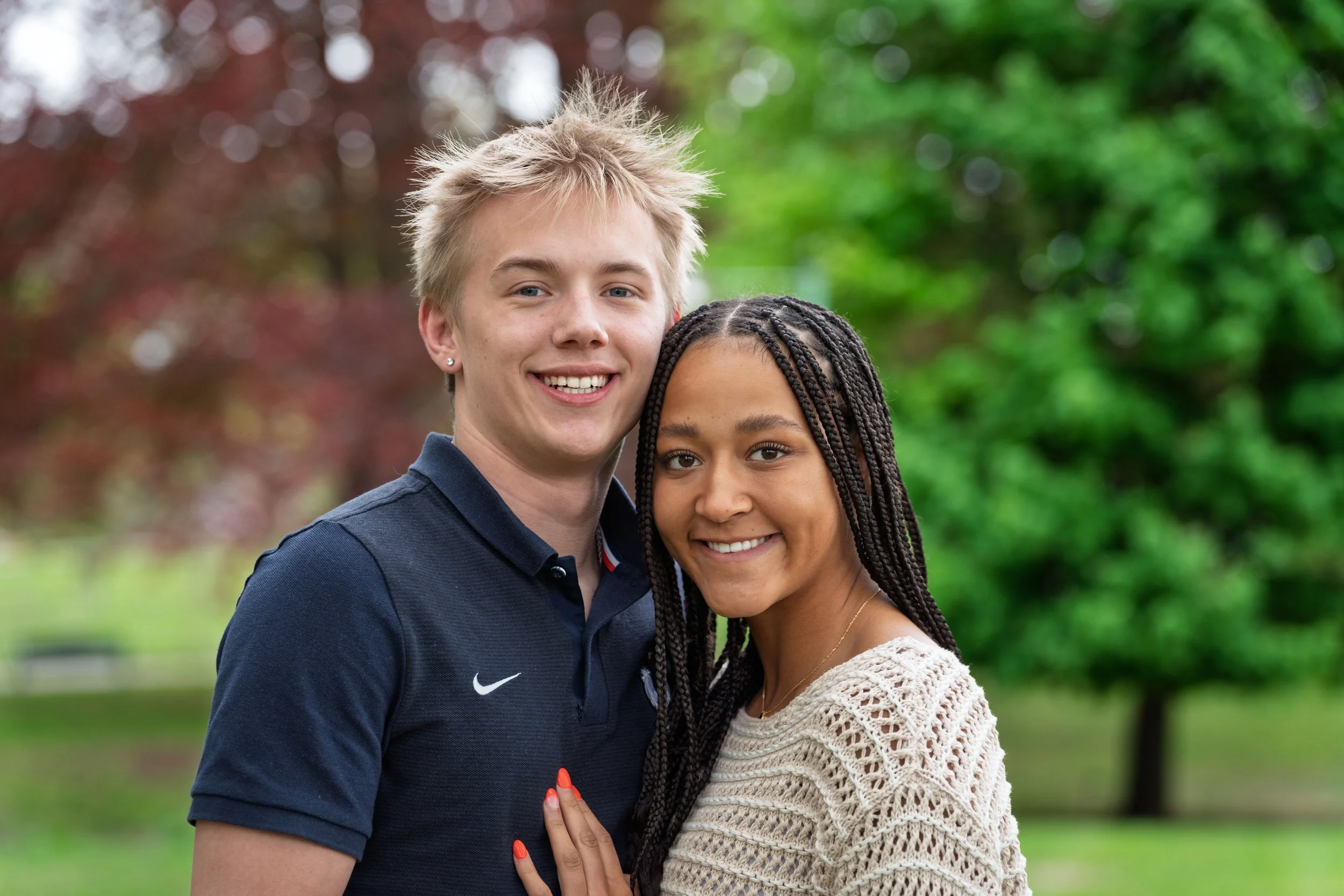 A young man and a young woman standing close together outdoors, smiling, with trees and greenery in the background.