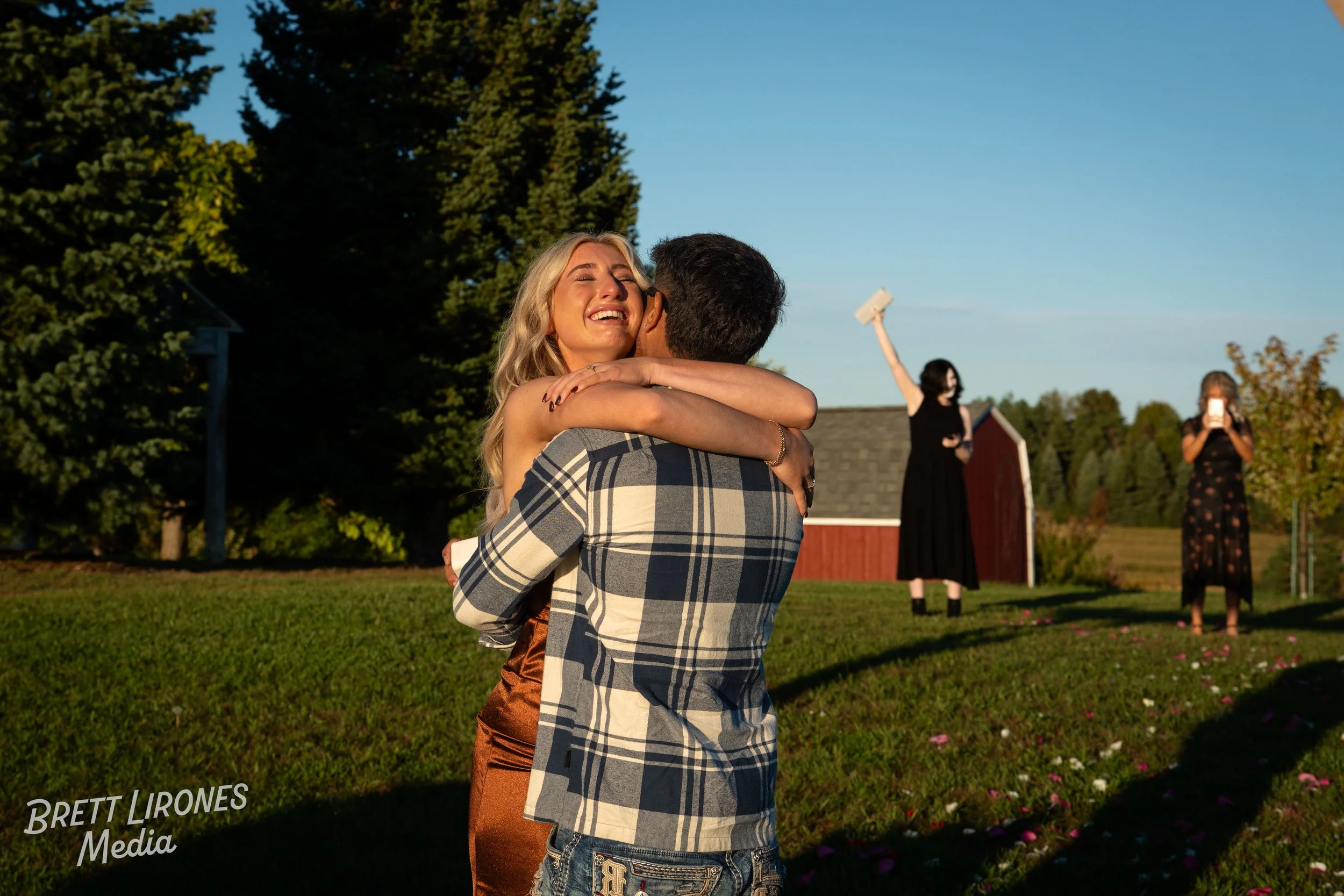 A couple hugging and smiling outdoors with a red barn and trees in the background, during sunset, with two women in black dresses taking photos in the distance.