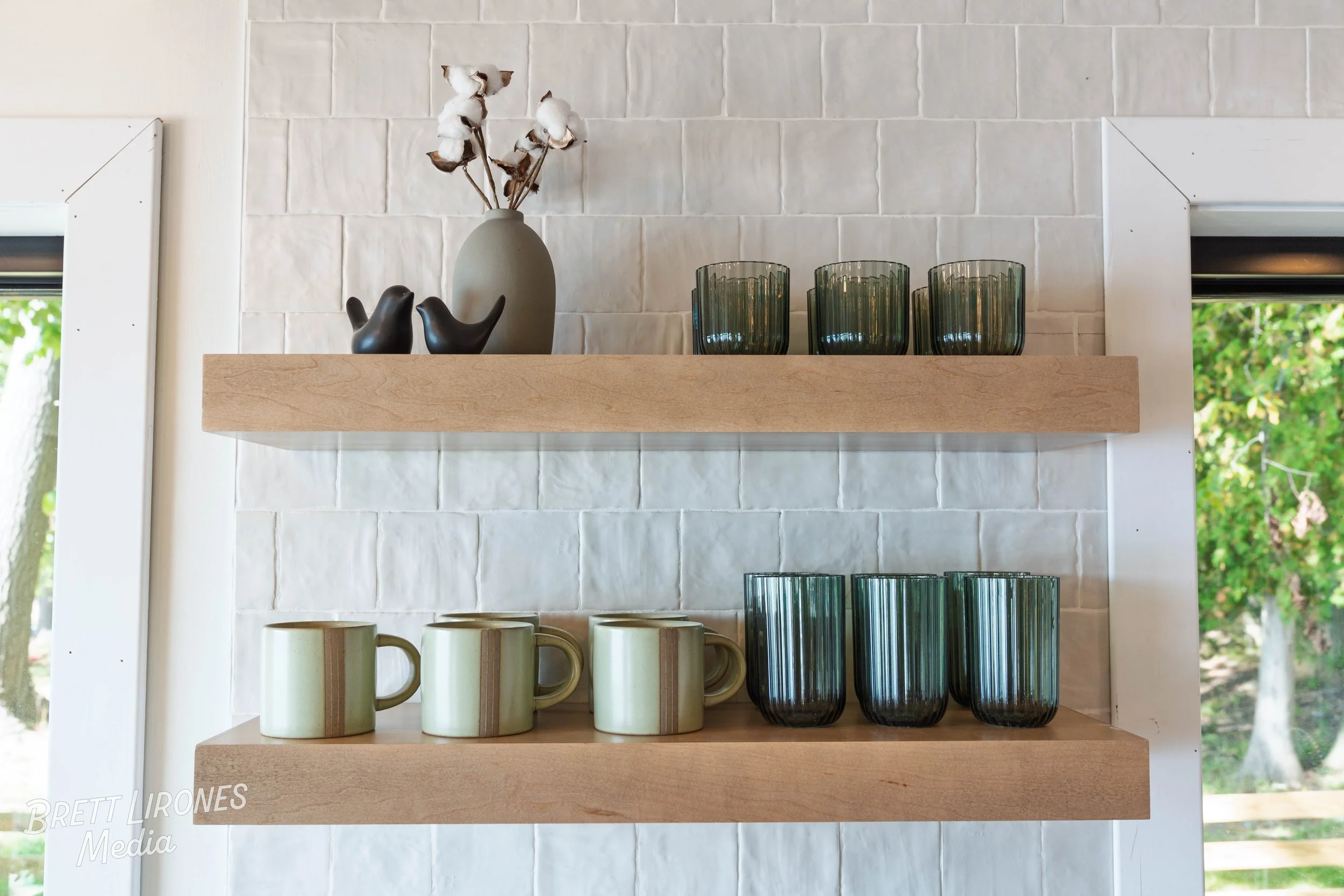 Two wooden shelves mounted on a white textured brick wall holding decorative items: a gray vase with dried cotton stems, black abstract ceramic sculptures, five green glass tumblers, and four beige ceramic mugs with a vertical stripe design.