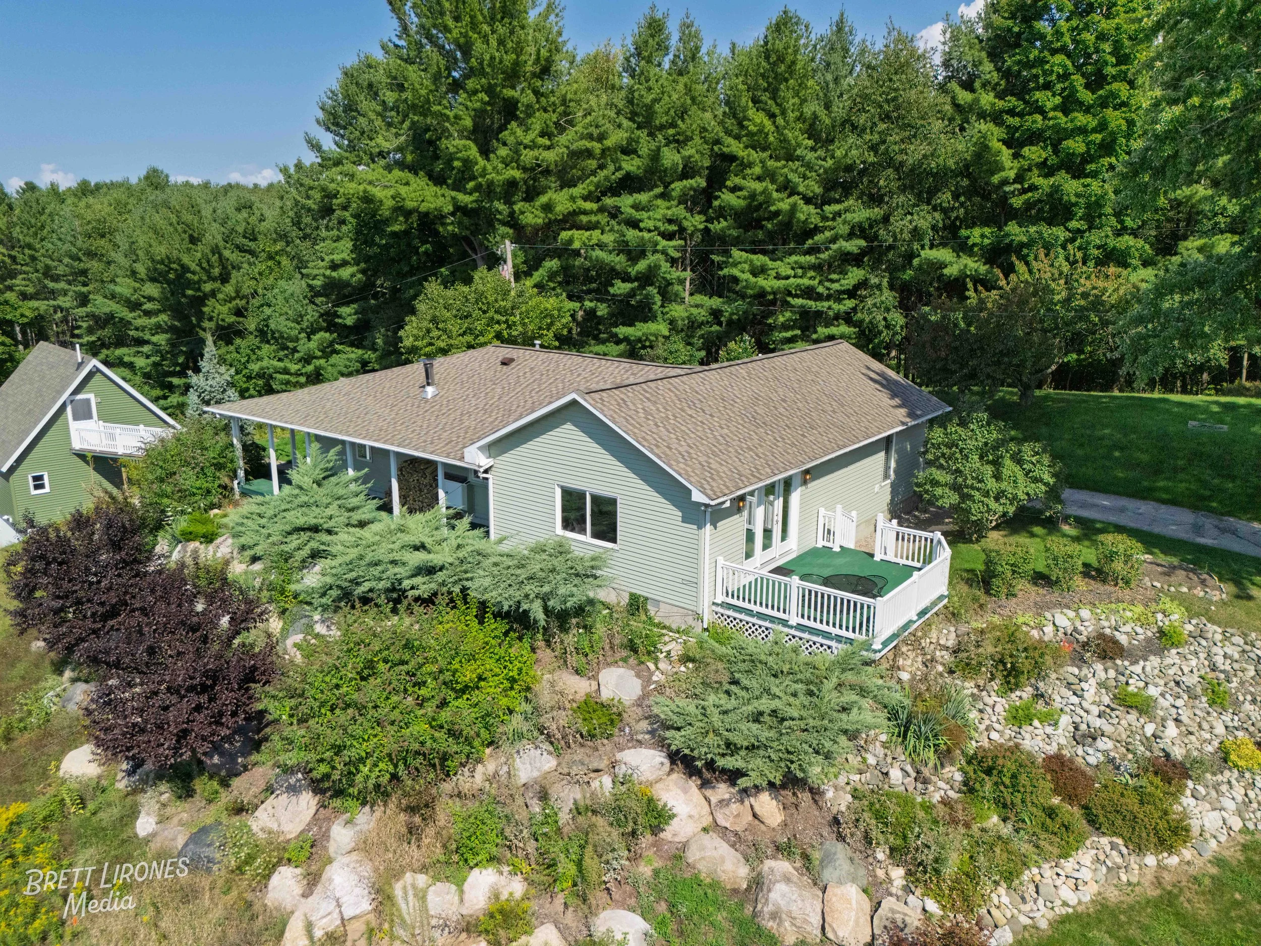 A light green house with a brown roof, a screened porch, and a deck with a hot tub, surrounded by greenery, trees, rocks, and shrubs, under a blue sky.