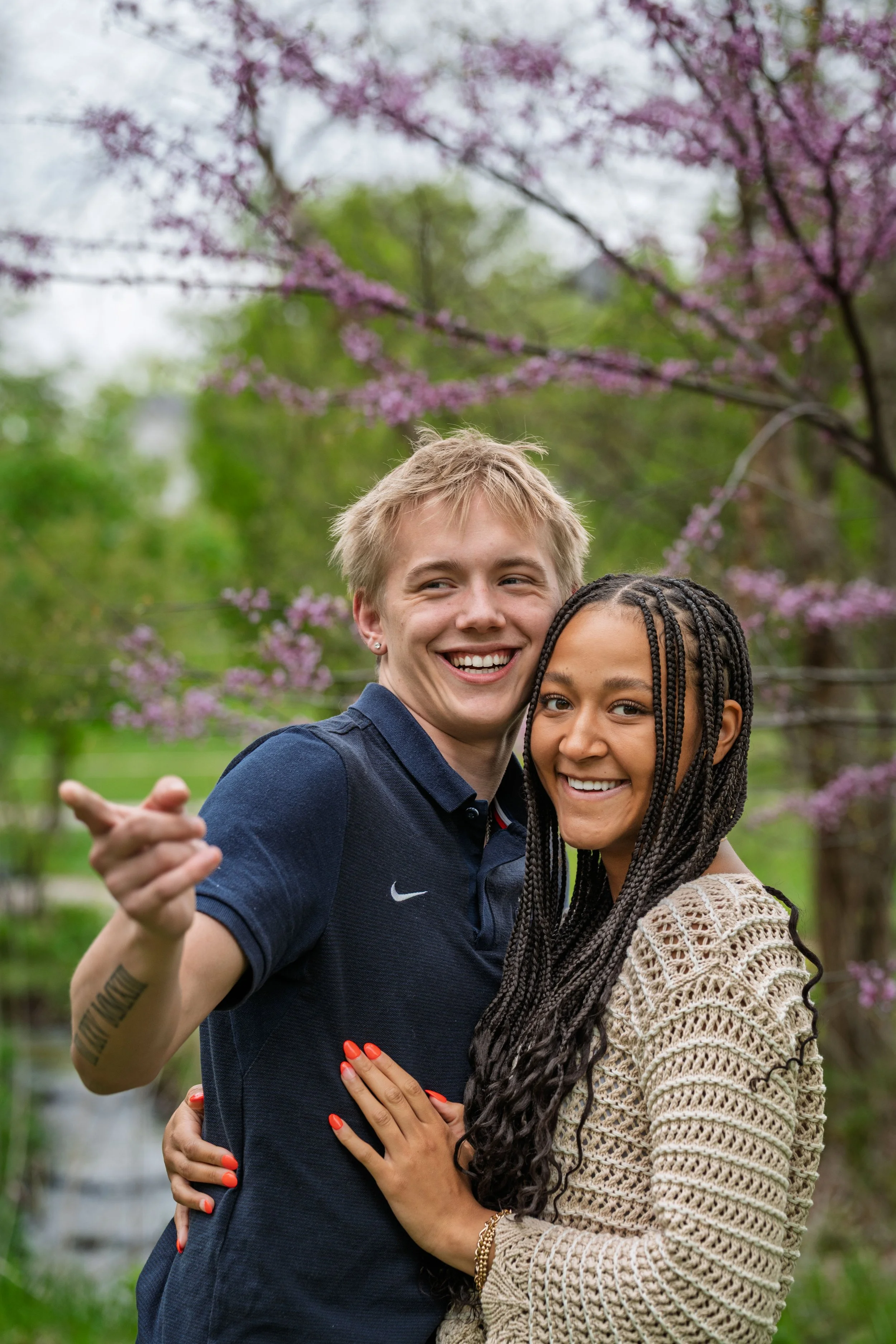 Two smiling young women hugging outdoors in a park with pink flowering trees in the background.