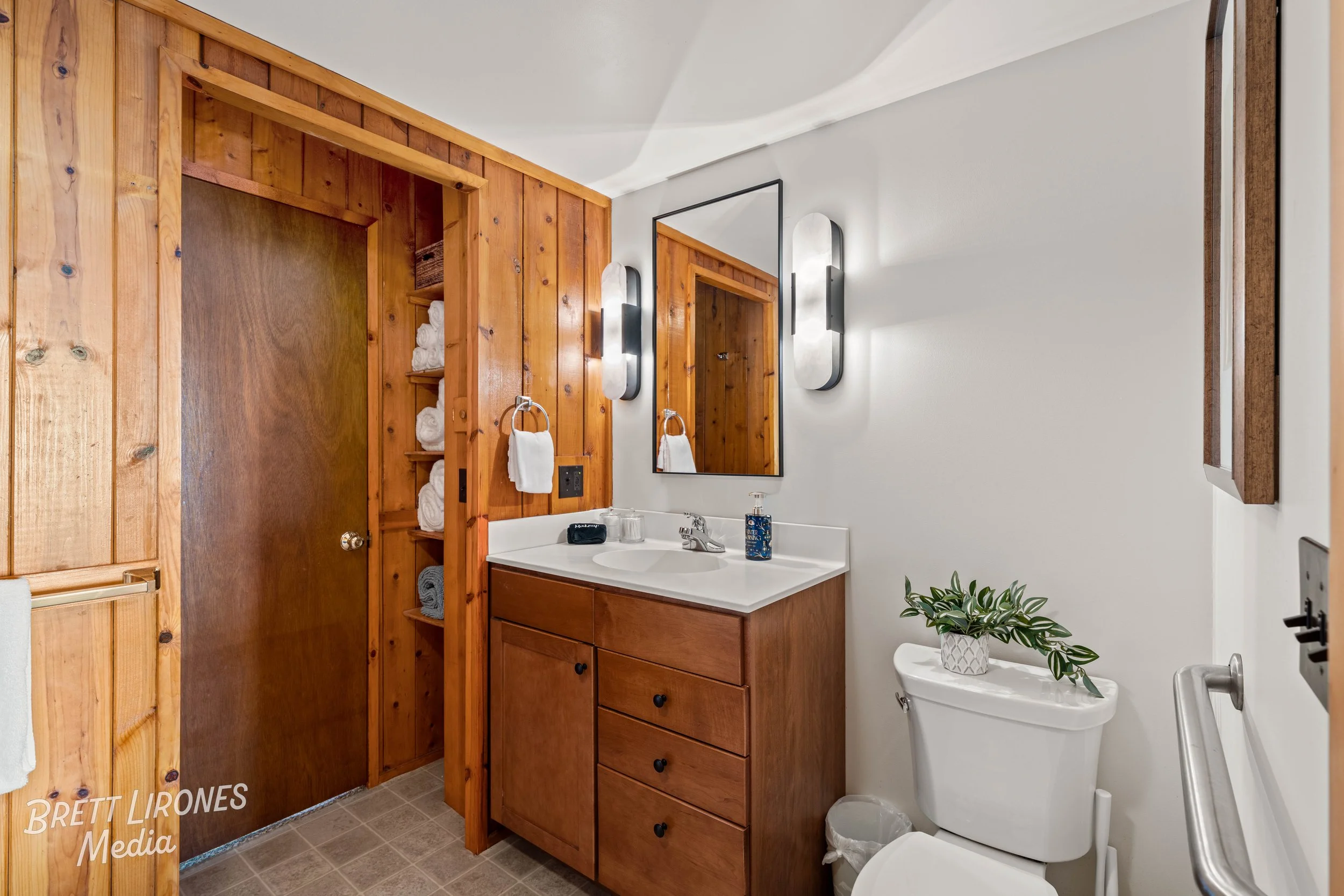 Bathroom with wooden accent walls, a mirror with two vertical light fixtures, a wooden vanity with a white countertop, a toilet with a potted plant on top, and towels on open shelves.