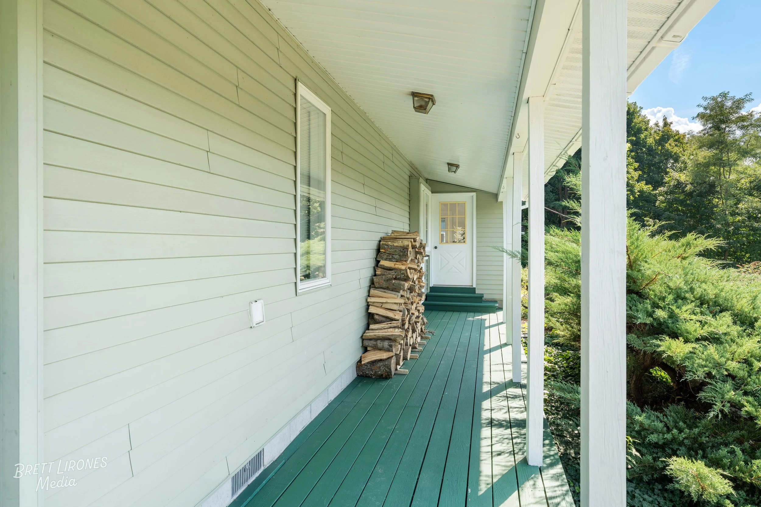 A front porch with green wooden flooring and light green siding, stacked firewood near the door, and lush greenery on the right side.