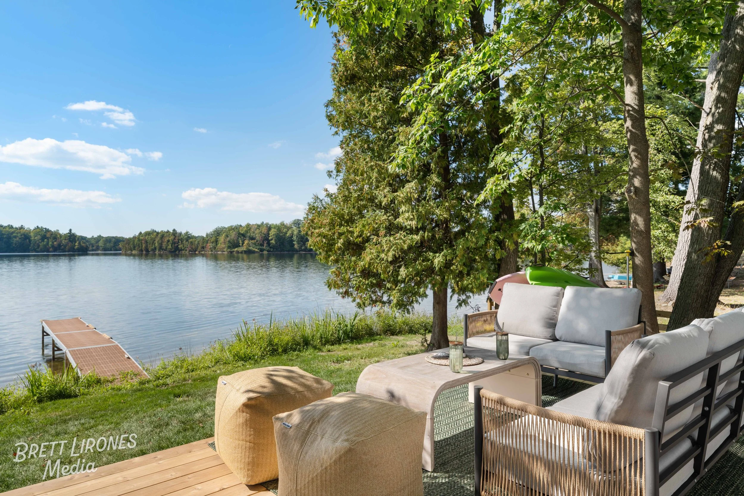 A lakeside outdoor patio with gray and beige furniture including a sofa, ottomans, and chairs, overlooking a calm lake with a foldable dock, surrounded by trees on a sunny day.
